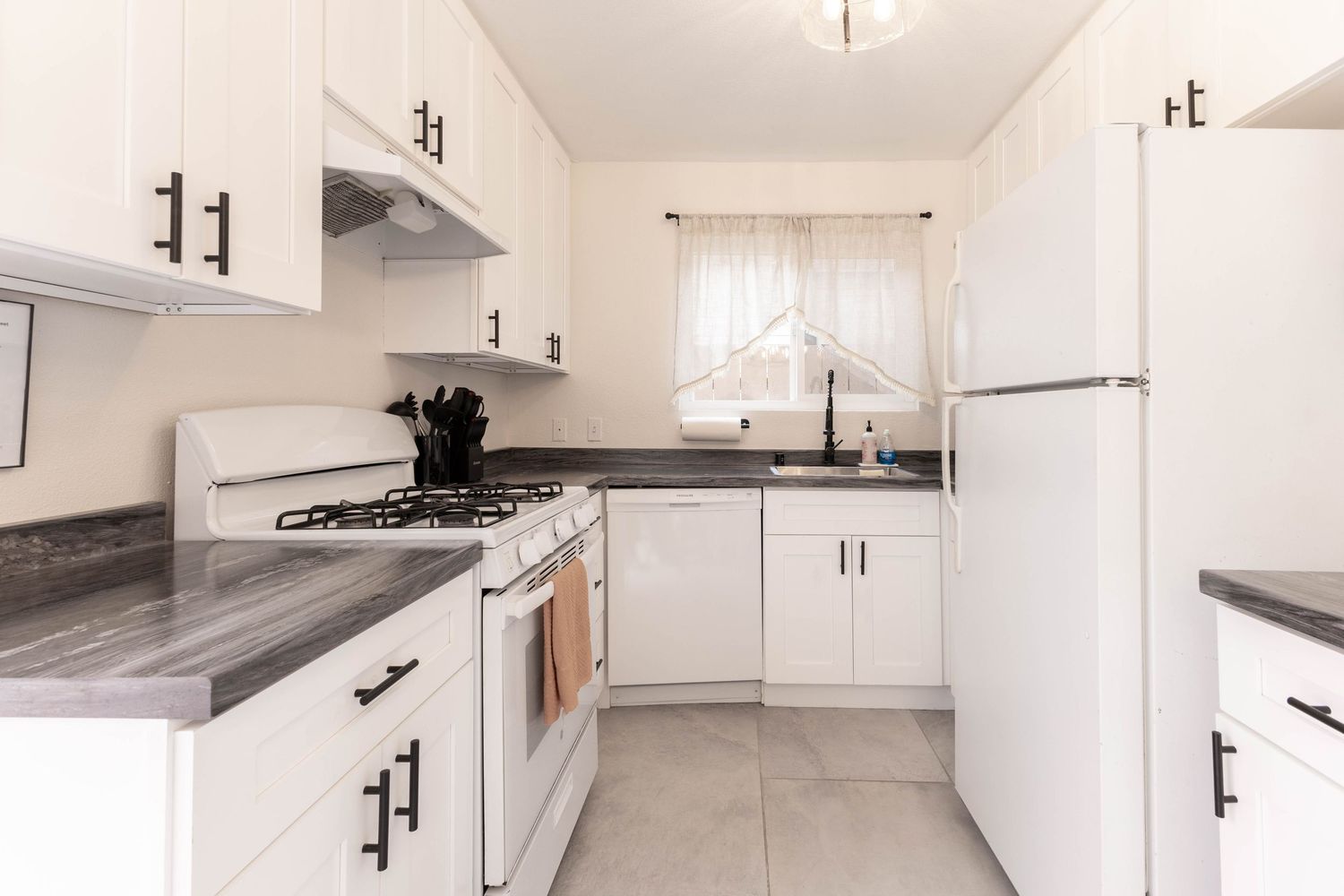 White kitchen with dark countertops, gas stove, refrigerator, black hardware, and tile flooring.