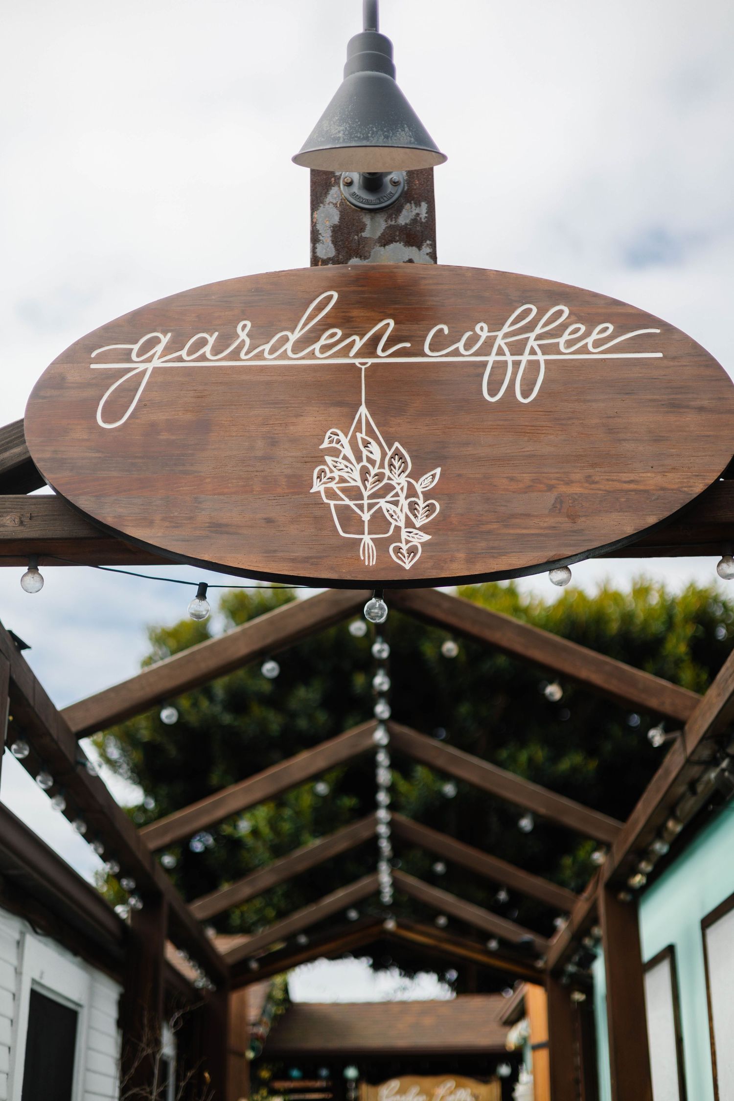 Rustic wooden round sign for Garden Coffee hanging under a pergola with string lights outdoors.