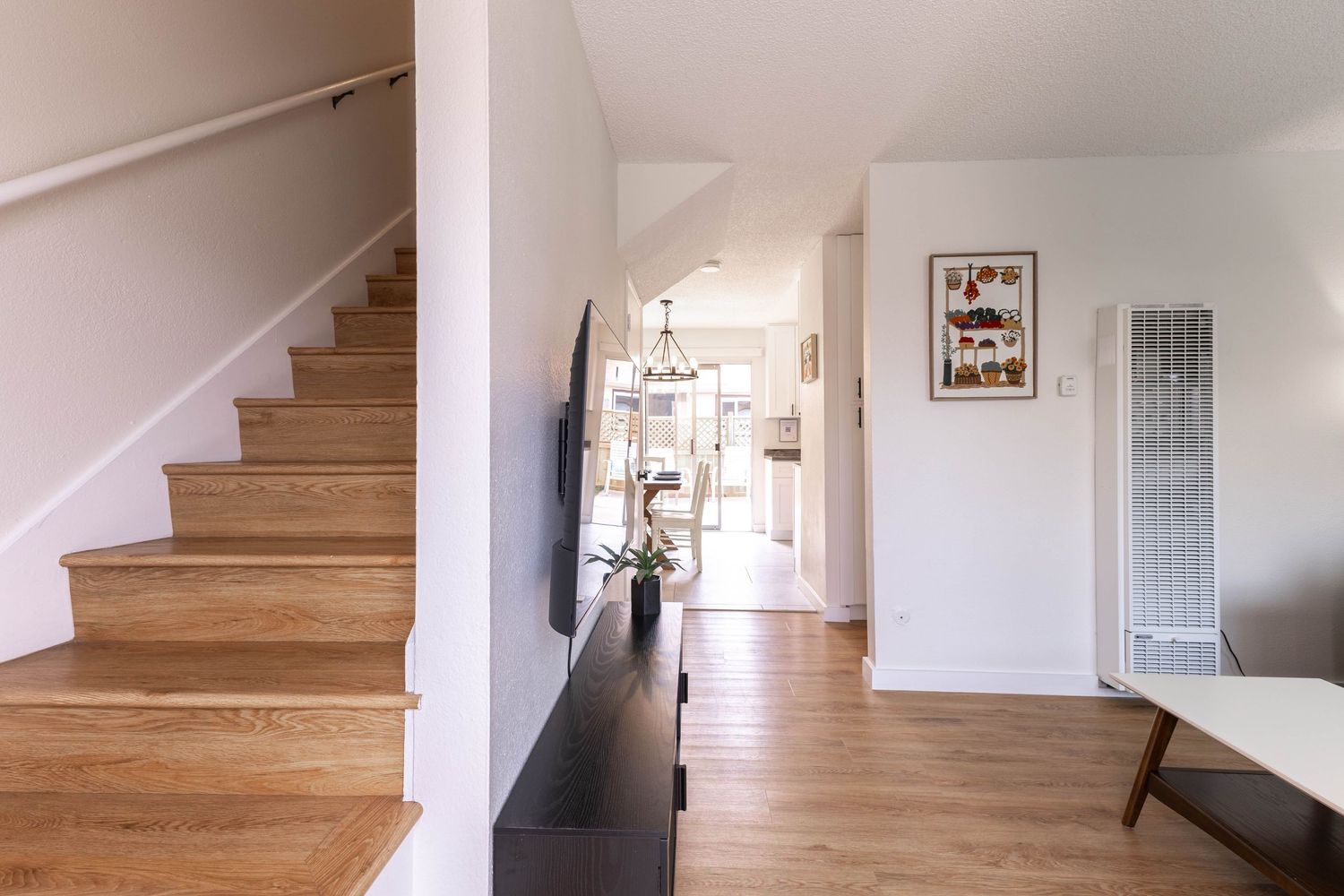 Modern open-plan living area with wooden staircase, hardwood floors, and bright hallway leading to kitchen.