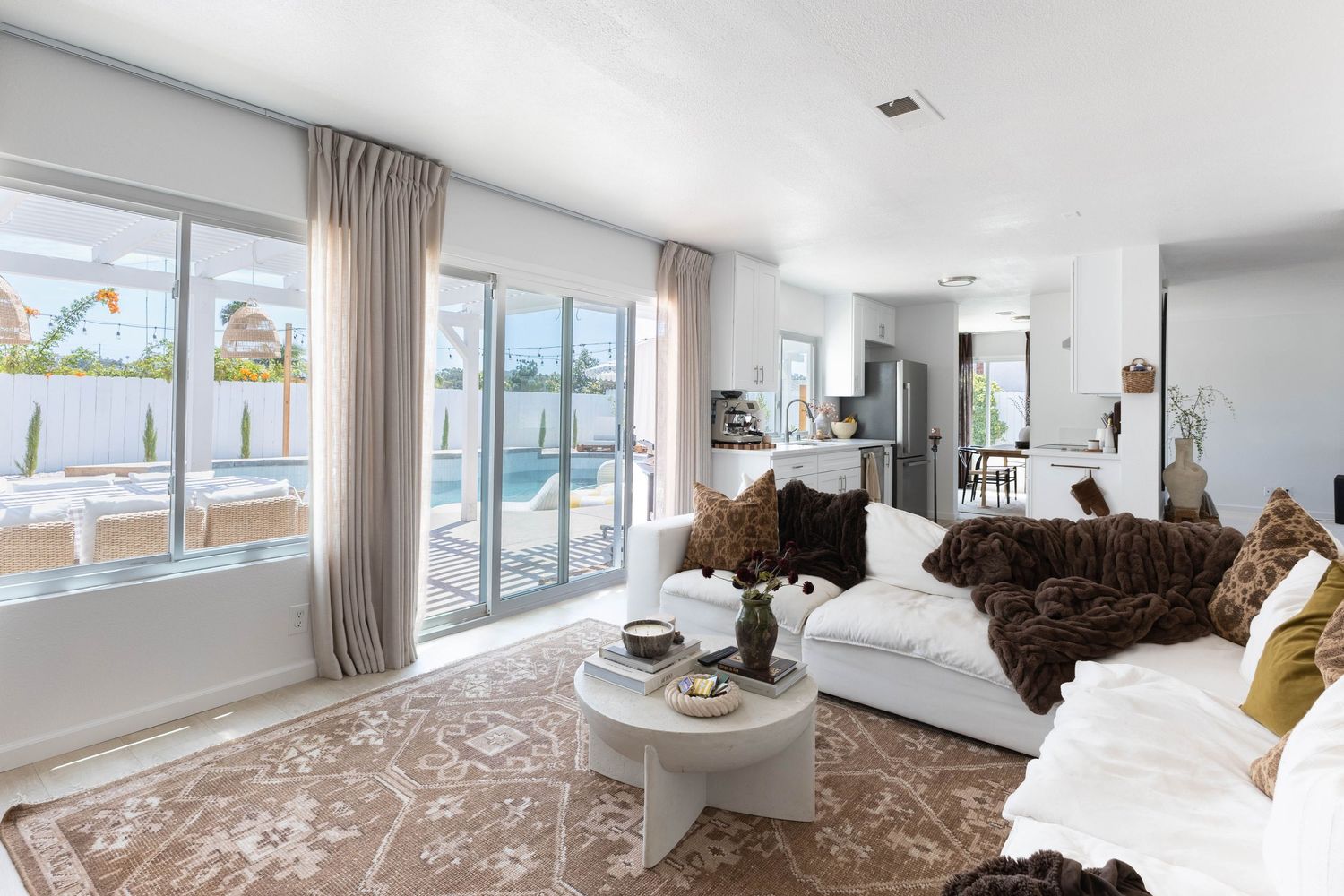 Bright living room with white sofas, fur throws, sliding glass doors, and pool view in a modern home.