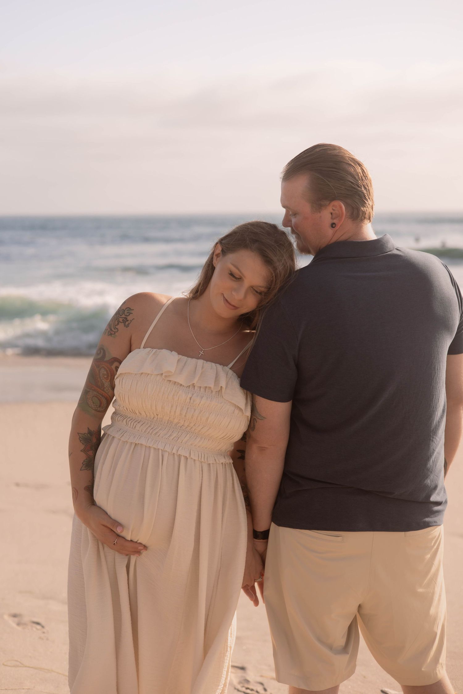 Pregnant couple sharing a tender moment on a sandy beach at sunset, woman in white dress cradling her bump.