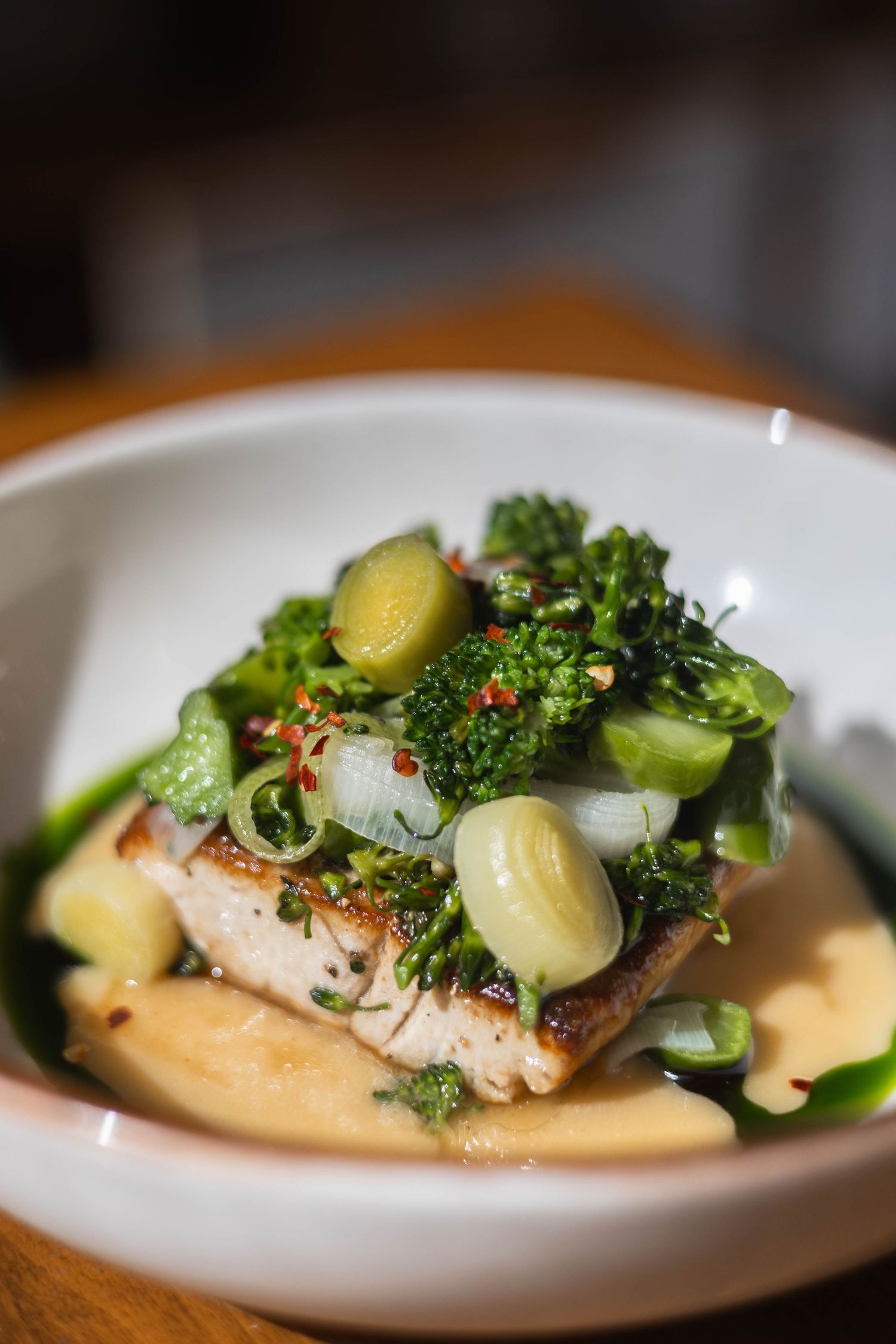 Seared fish fillet served in a white bowl with broccoli, green onions, olives, and creamy sauce.