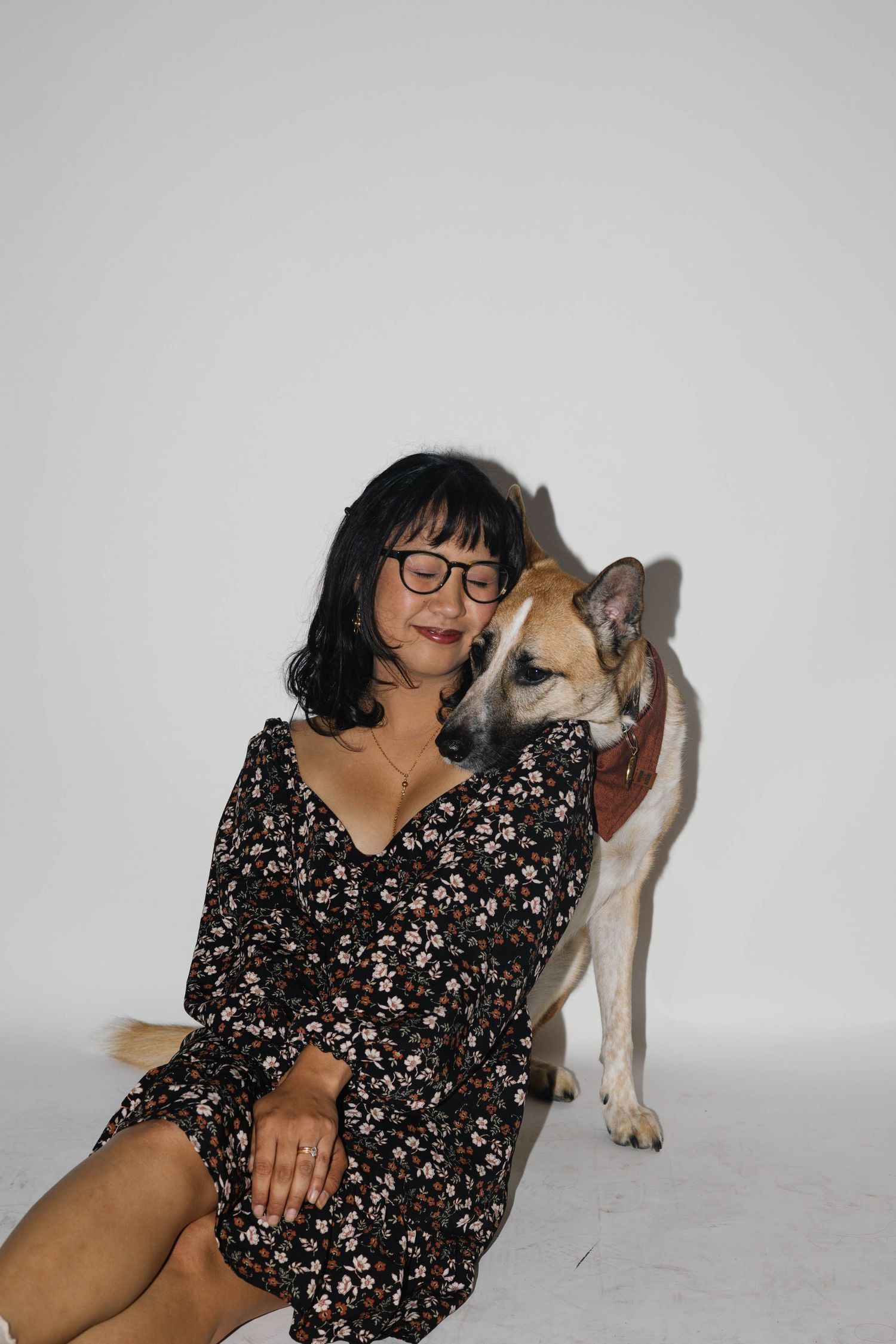 Woman in floral dress sitting with a tan dog nuzzling her cheek against a white background.