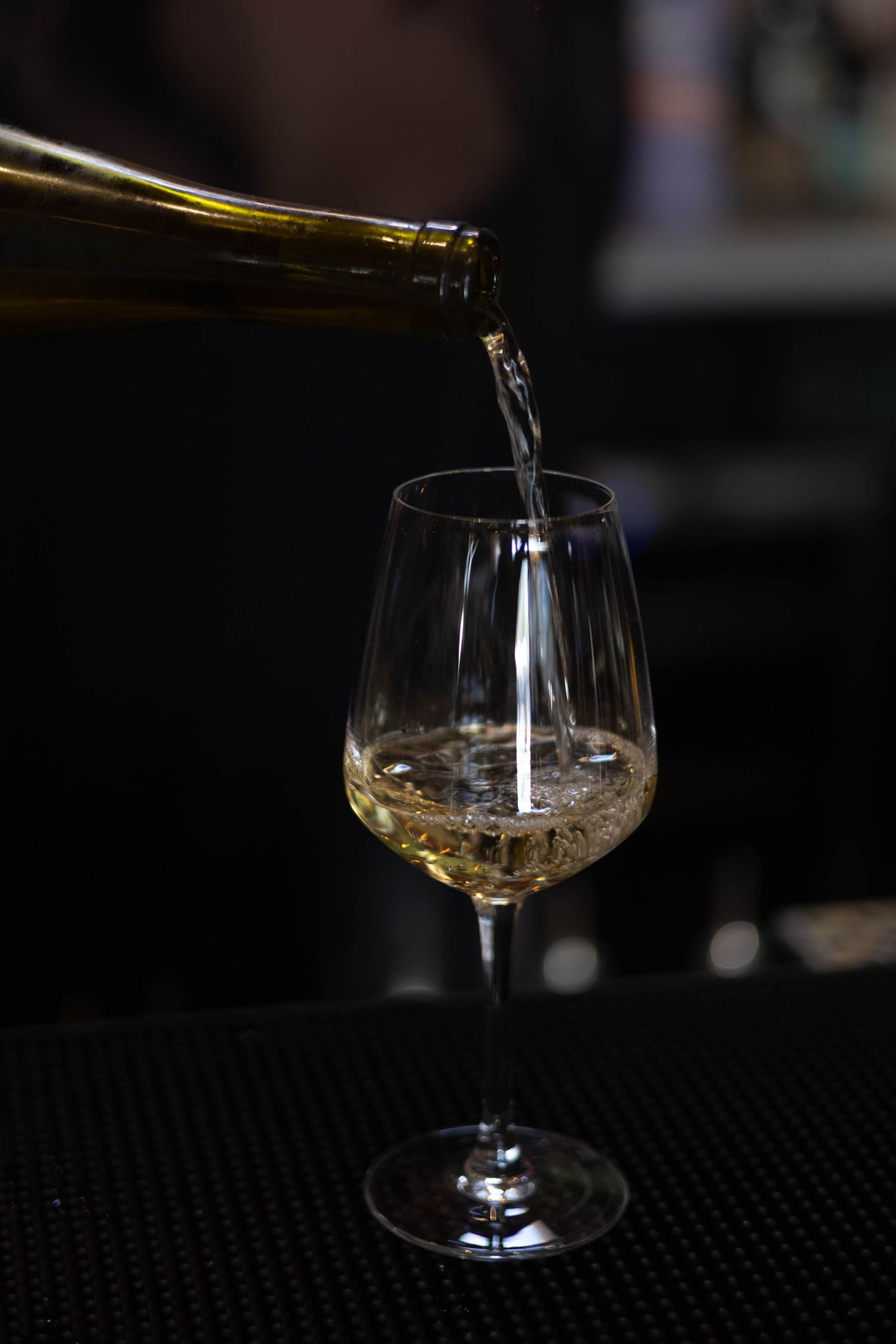 White wine being poured from a green bottle into an elegant crystal wine glass on a dark bar surface.
