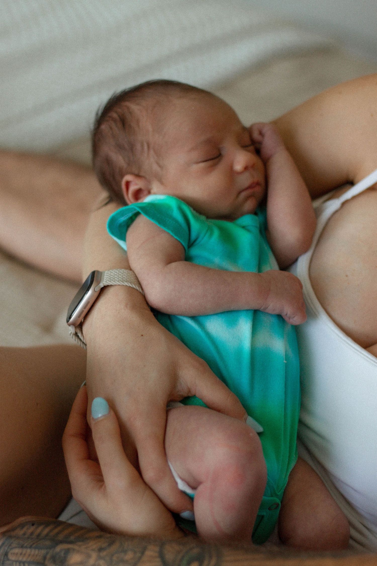 Newborn baby in teal onesie sleeping peacefully in mother's arms, with parent wearing an Apple Watch.