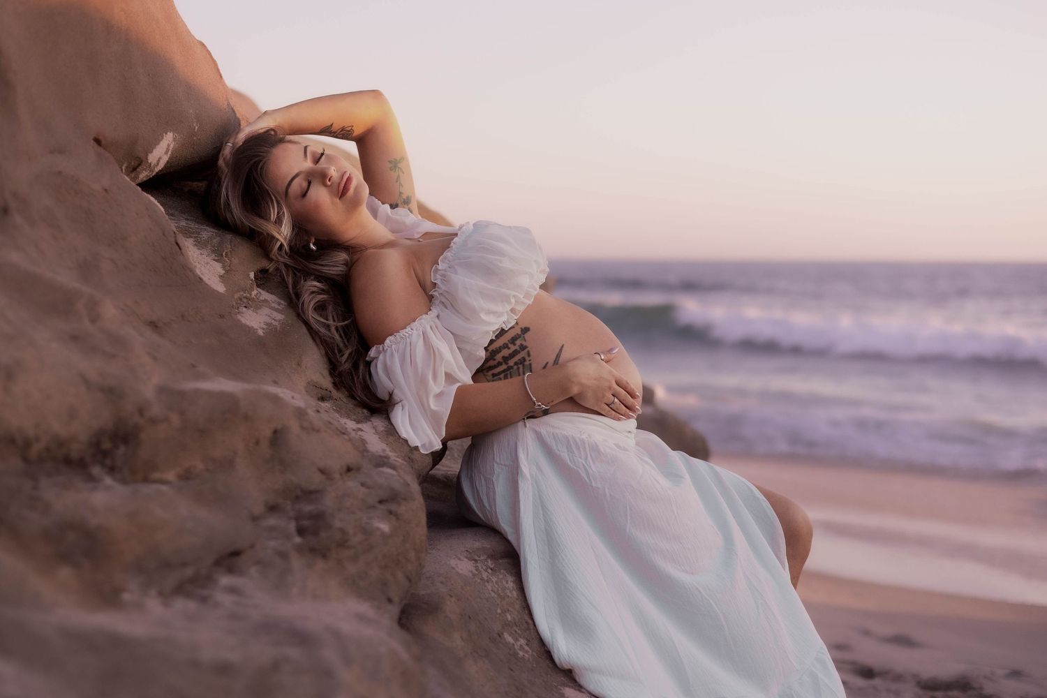 Pregnant woman in white off-shoulder dress leaning against beach rocks at golden hour sunset.