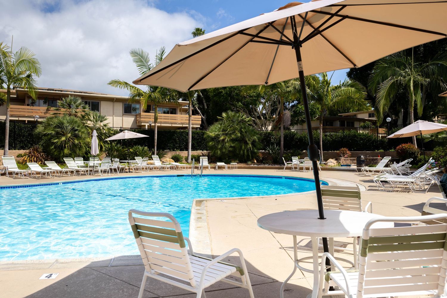 Outdoor hotel pool with white patio furniture, large tan umbrellas, and lush palm trees on a sunny day.