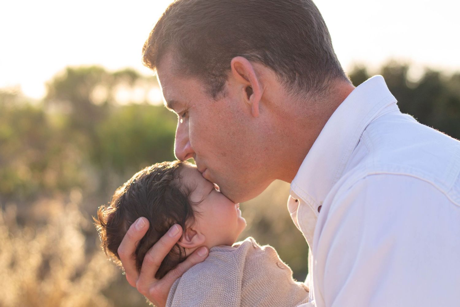 Loving father gently kissing newborn baby outdoors in soft golden natural light.