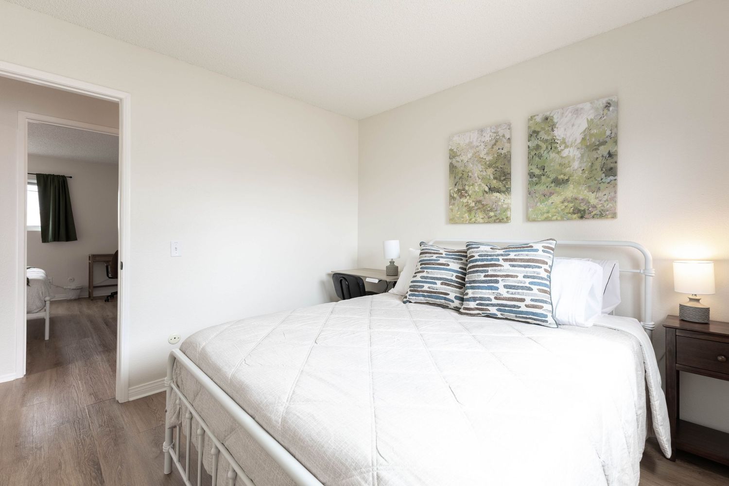Minimalist white bedroom with queen bed, blue pillows, nightstand lamp, and nature wall art.