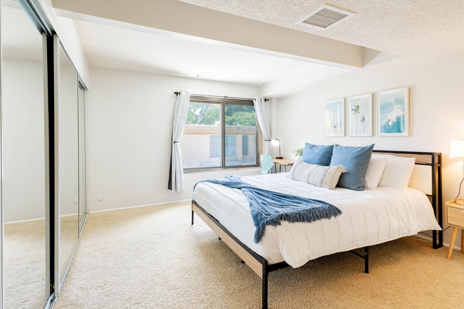 Bright bedroom with a queen bed, blue accent pillows, mirrored closet doors, and a large window with natural light.