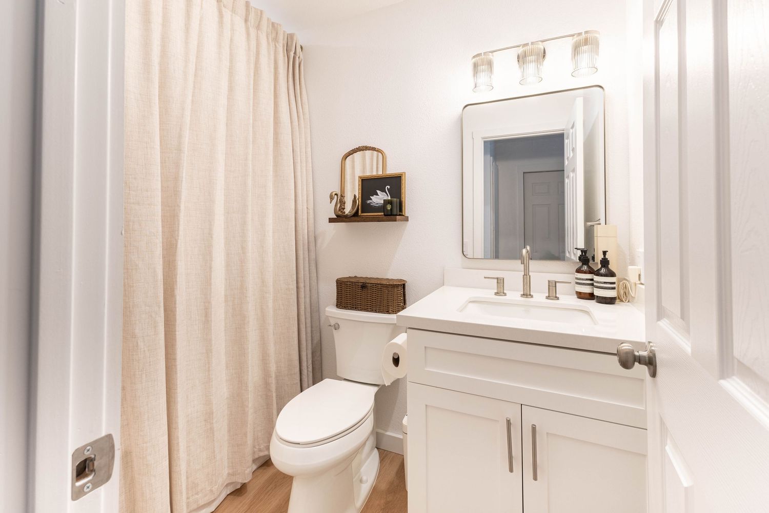 A bright, clean bathroom featuring a white vanity, framed mirror, toilet, and neutral shower curtain with wood-look flooring.