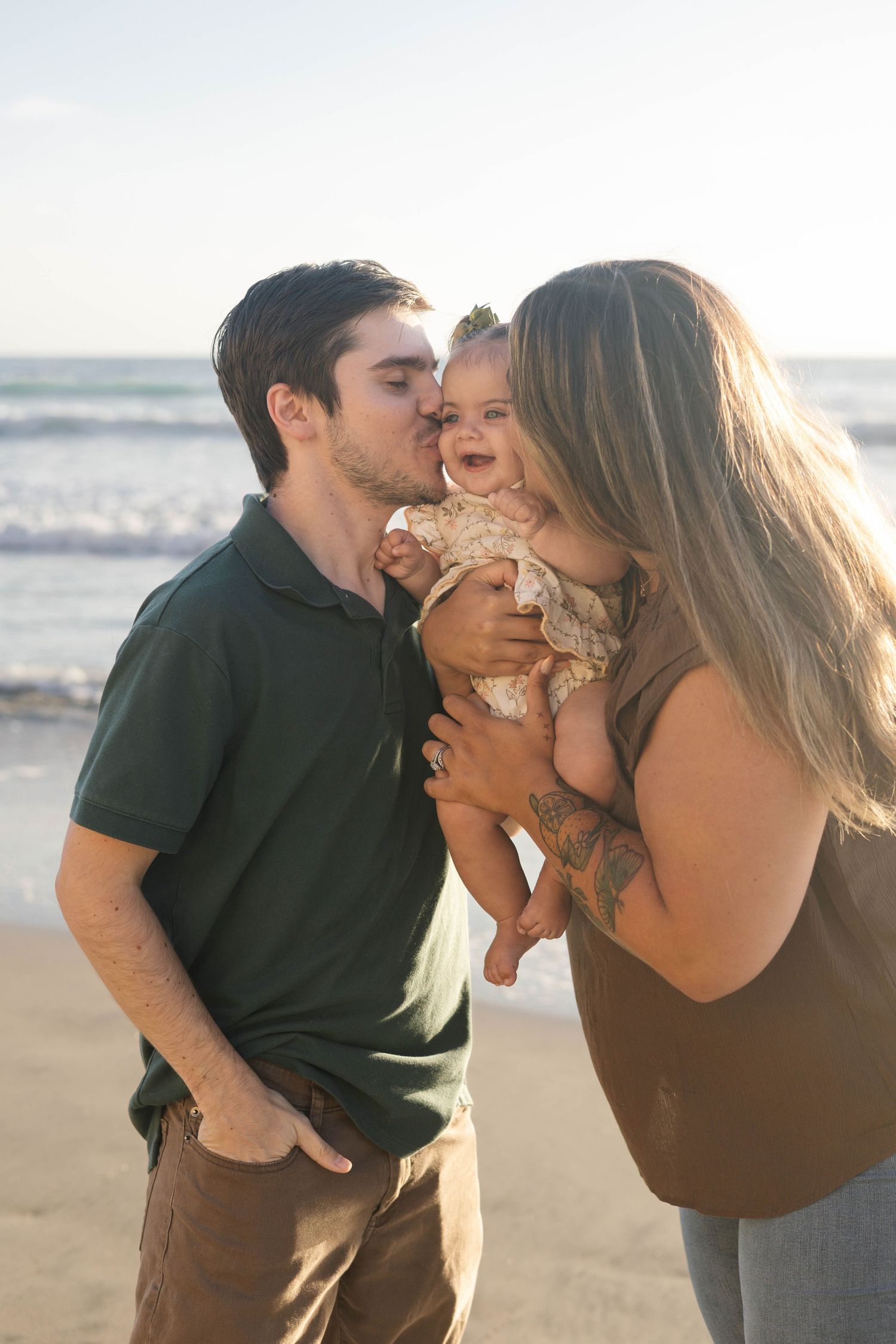 Young couple kissing their smiling baby on a sunny beach, creating a heartwarming family moment by the ocean.