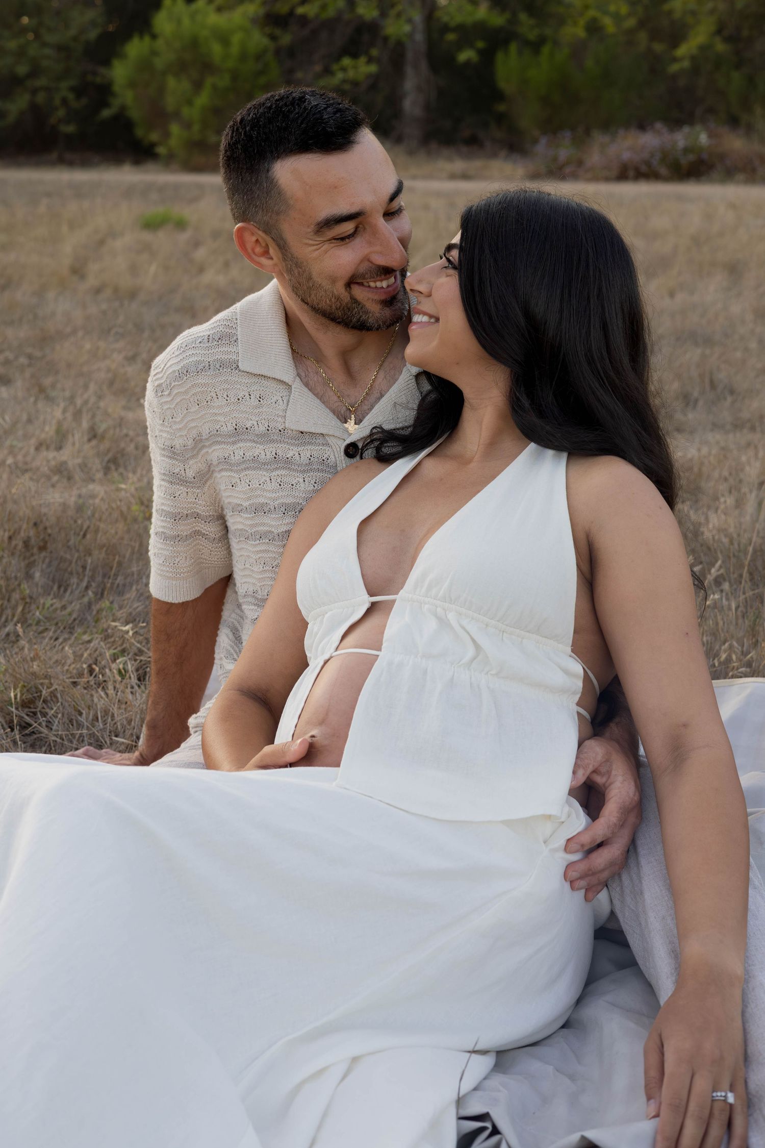 A couple shares a tender moment outdoors, with a pregnant woman in a white dress and her partner smiling at each other.