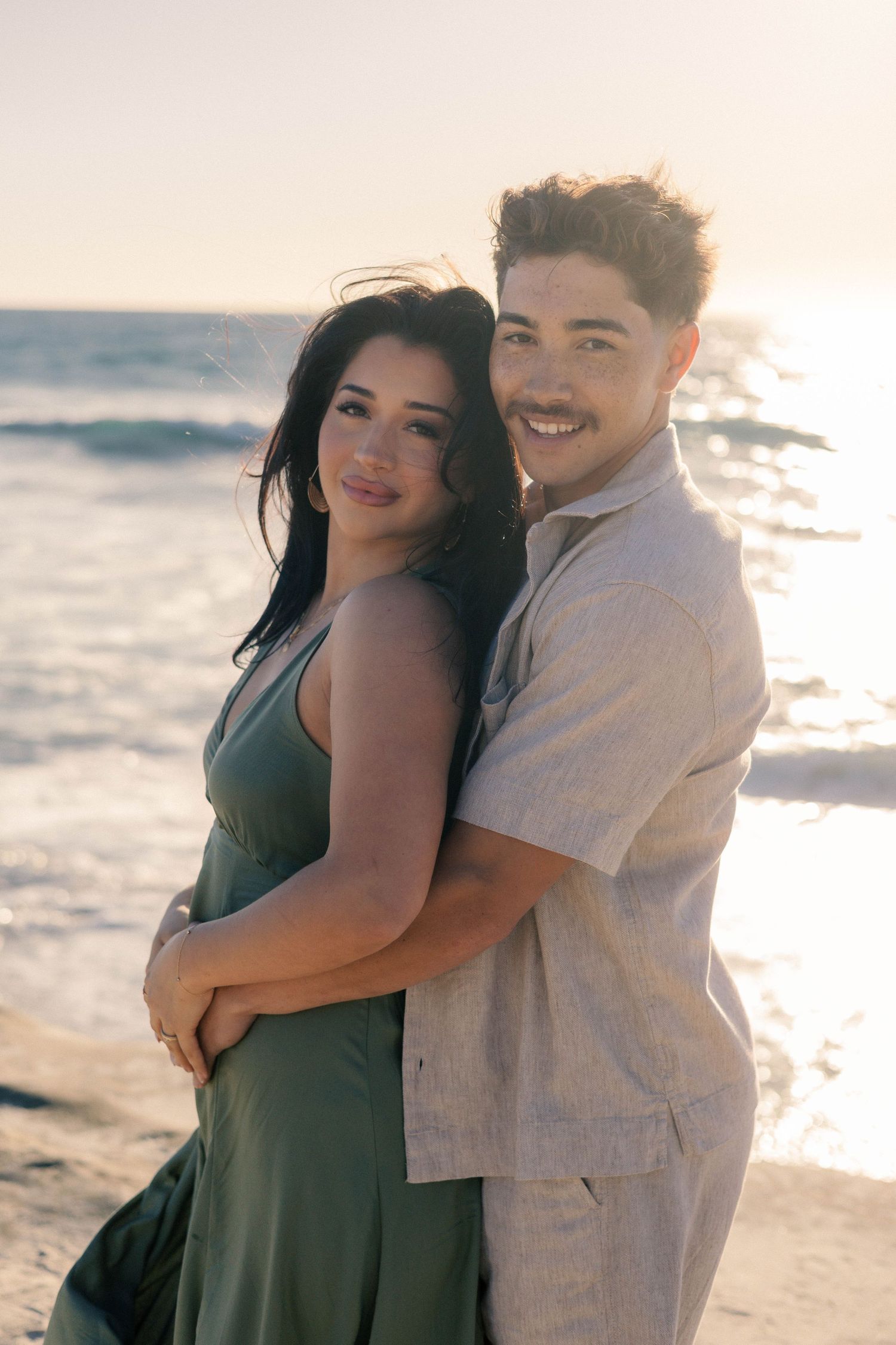 Smiling couple hugging on a sunlit beach, man in linen shirt embracing woman in green dress.
