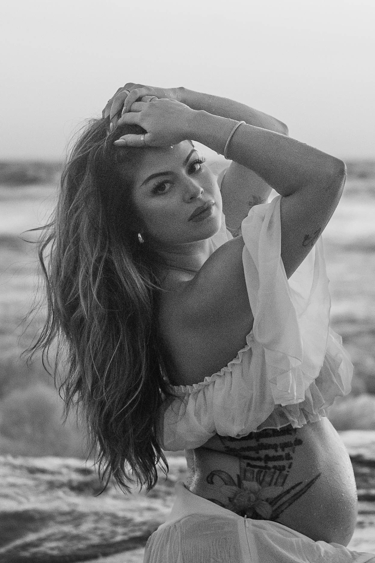 Black and white portrait of pregnant woman in white off-shoulder top posing on beach with hands in hair.