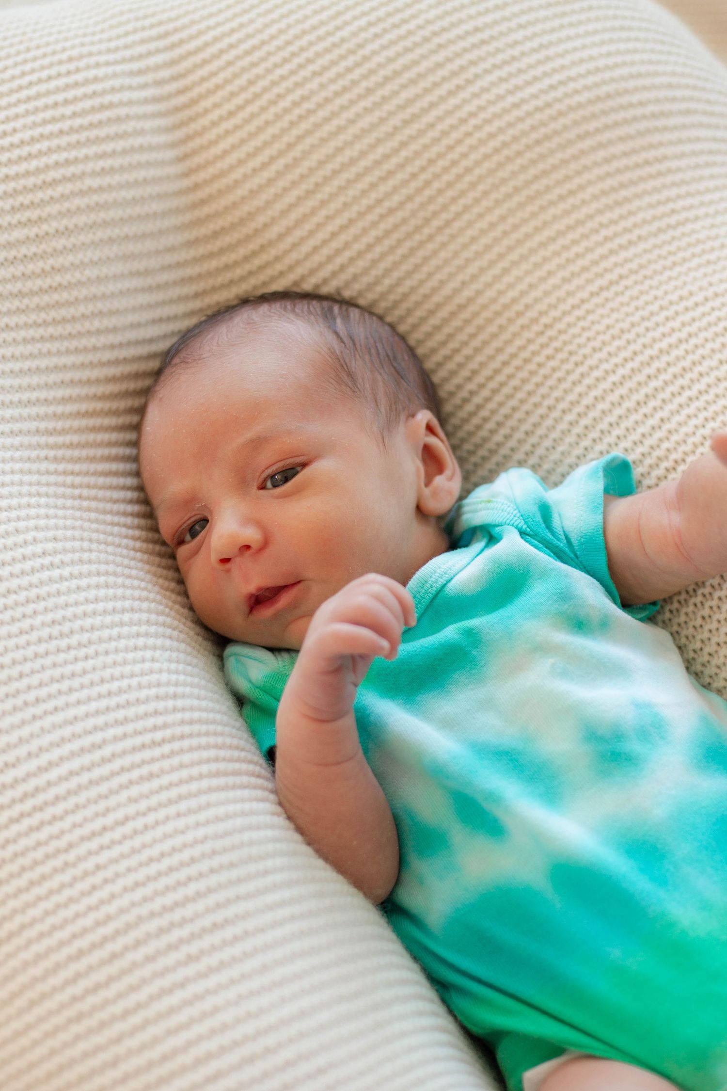 Newborn baby wearing turquoise outfit lying on beige textured pillow looking at camera.