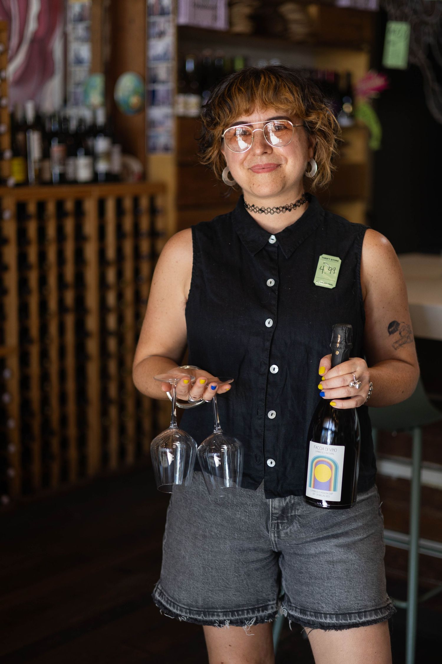 A smiling woman in glasses holds wine glasses and a bottle in a cozy wine shop interior.
