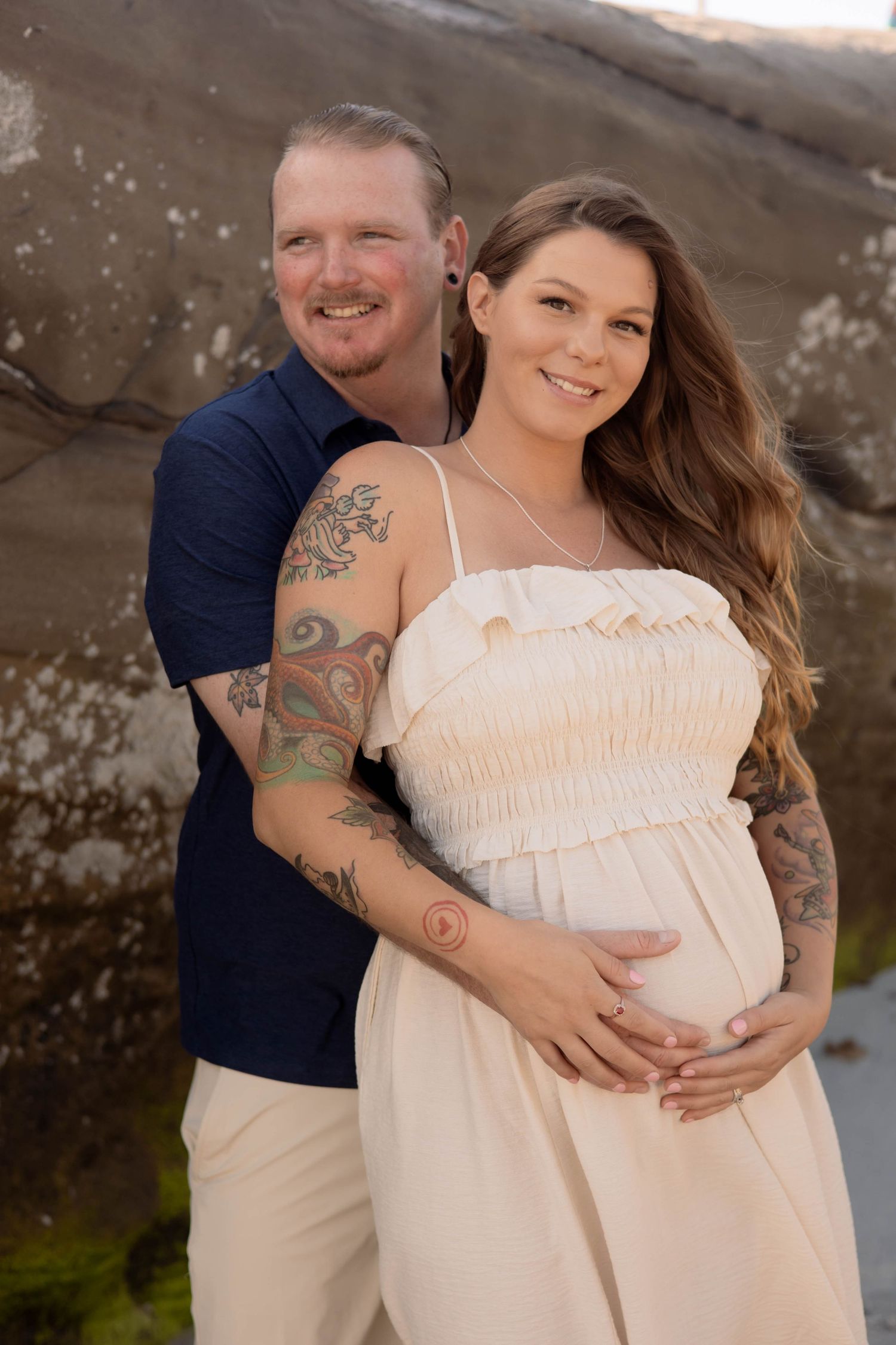A tattooed couple poses outdoors for a maternity photo, the woman in a white dress cradling her baby bump.