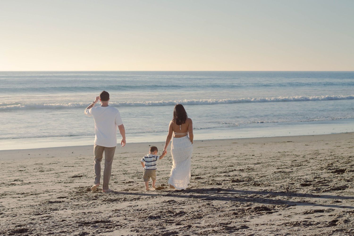A family of three walks toward the ocean on a peaceful sandy beach during sunset.