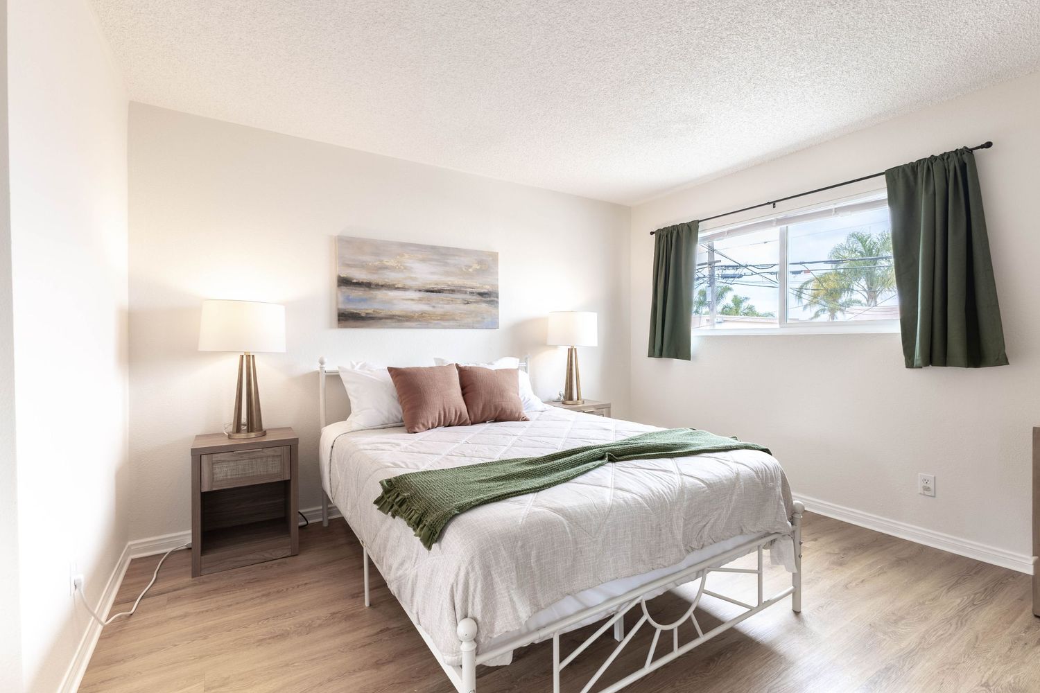 Bright bedroom with white metal bed frame, green throw blanket, pink pillows, wood floors, and green curtains by a sunlit window.