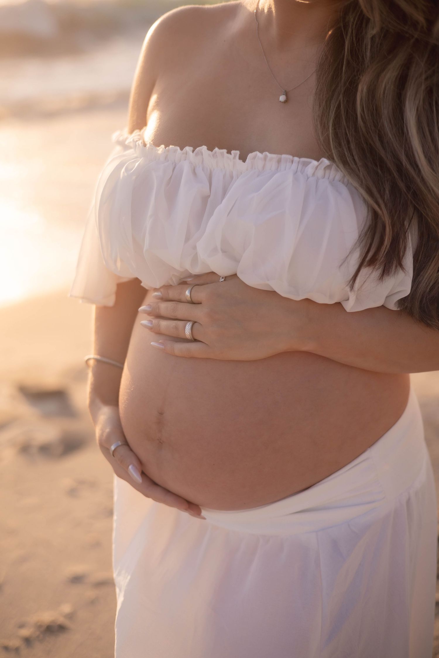 Close-up of pregnant woman in white off-shoulder top gently holding bare baby bump at beach.