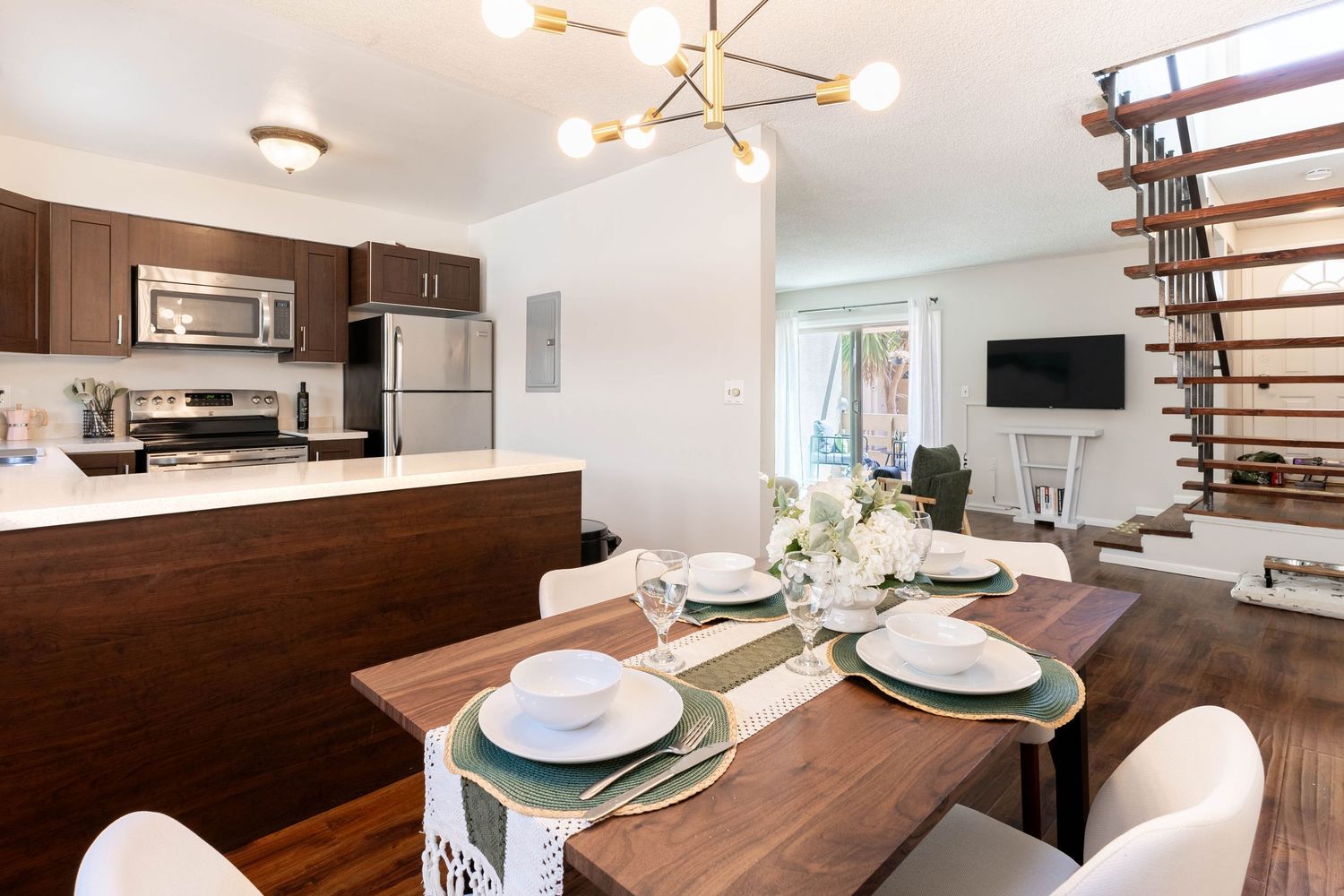 Modern open-plan dining and kitchen area with wooden table, white chairs, stainless appliances, and stylish sputnik chandelier.