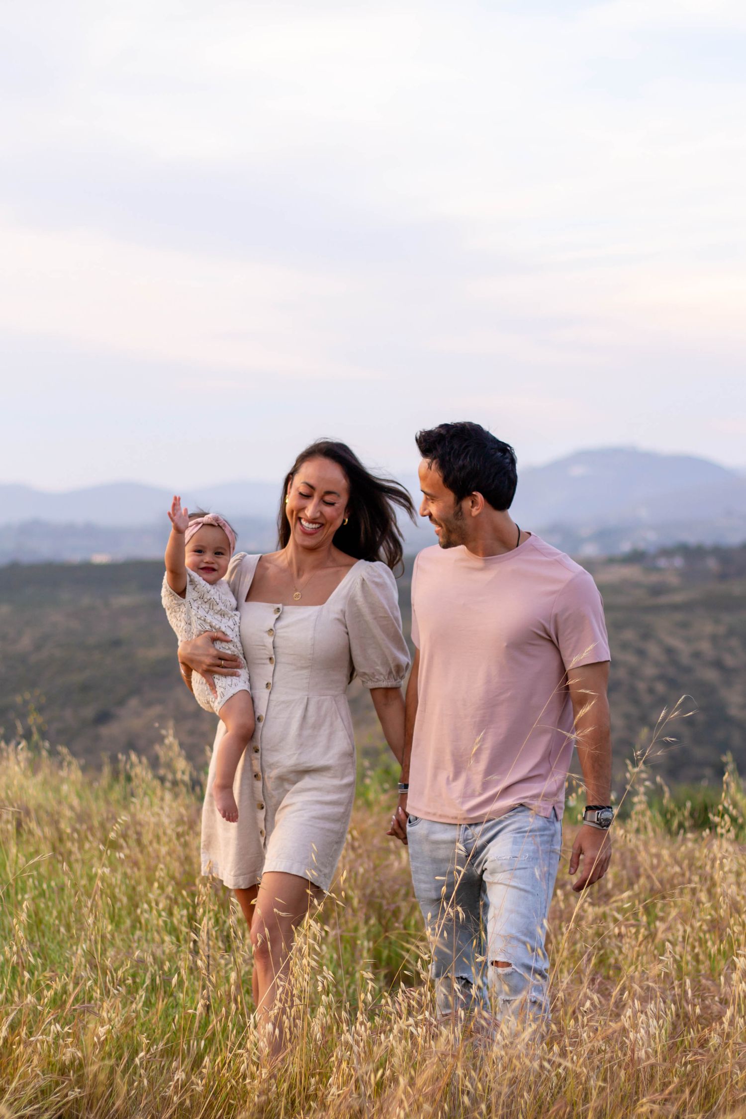 Happy young family walking through tall grass meadow with baby girl at golden hour.