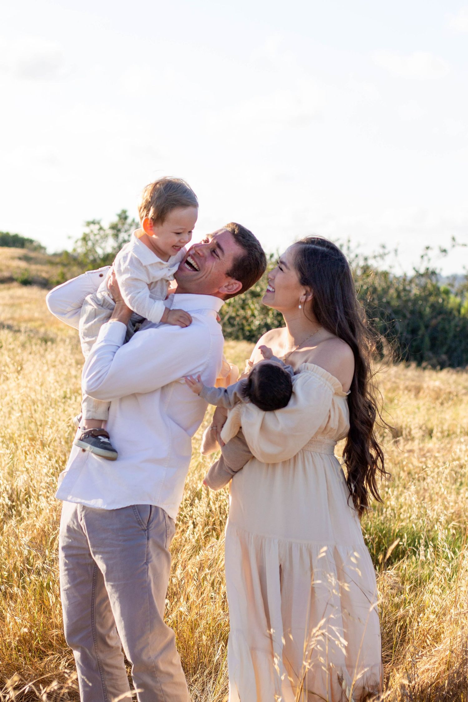 Joyful father lifts laughing toddler while mother holds newborn in sunny golden grass field.