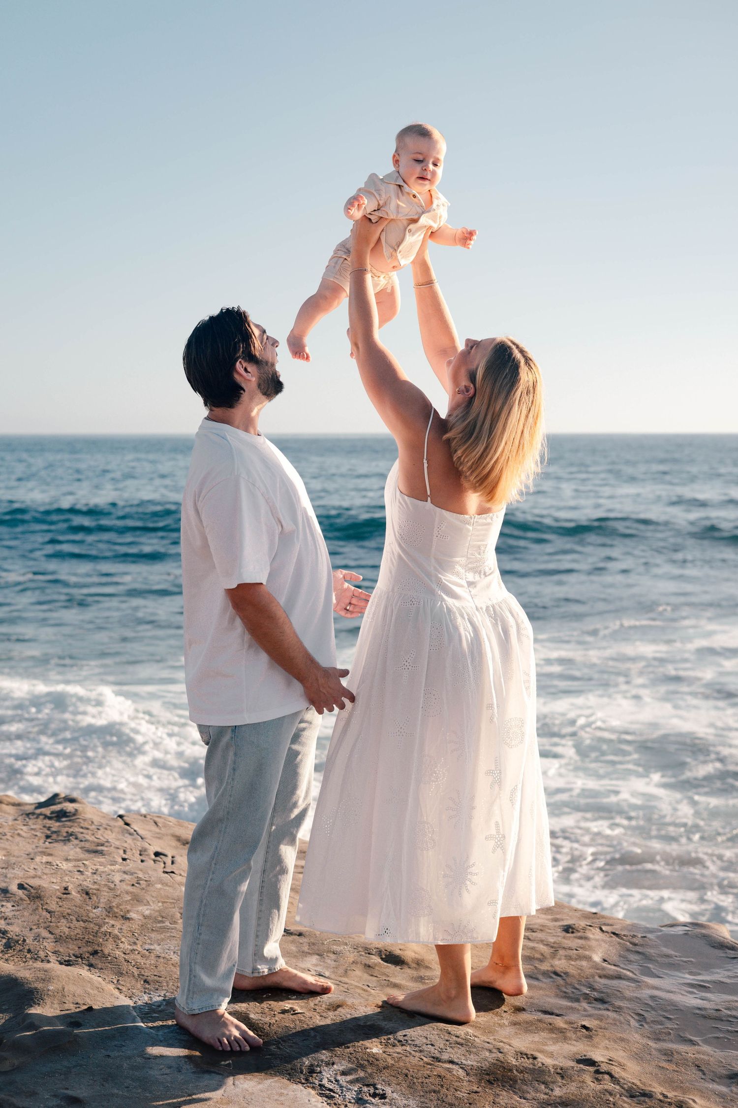 Happy family with mother lifting baby in the air on rocky beach, father watching, ocean backdrop.