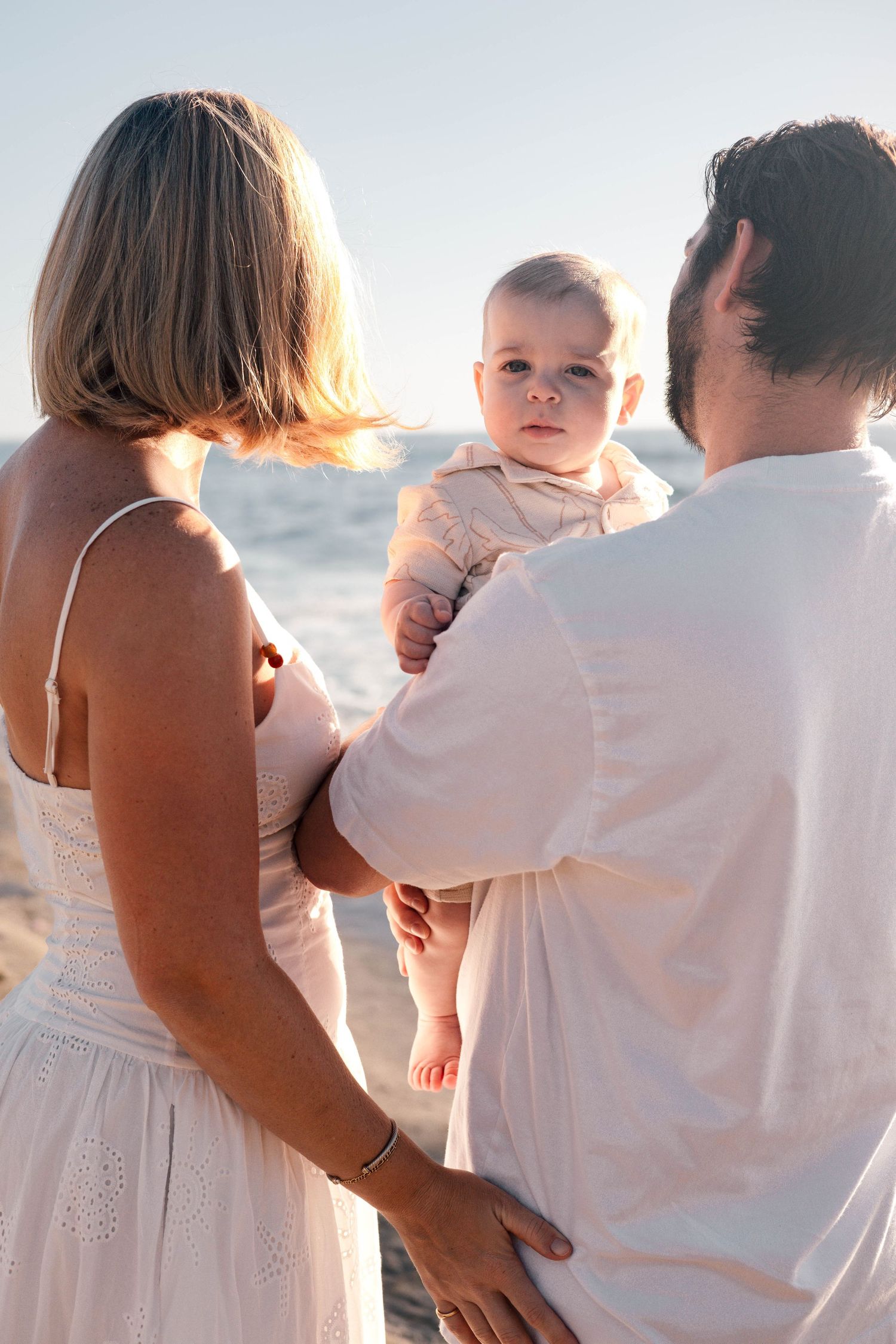 A couple holding their baby at the beach during golden hour, with the ocean in the background.