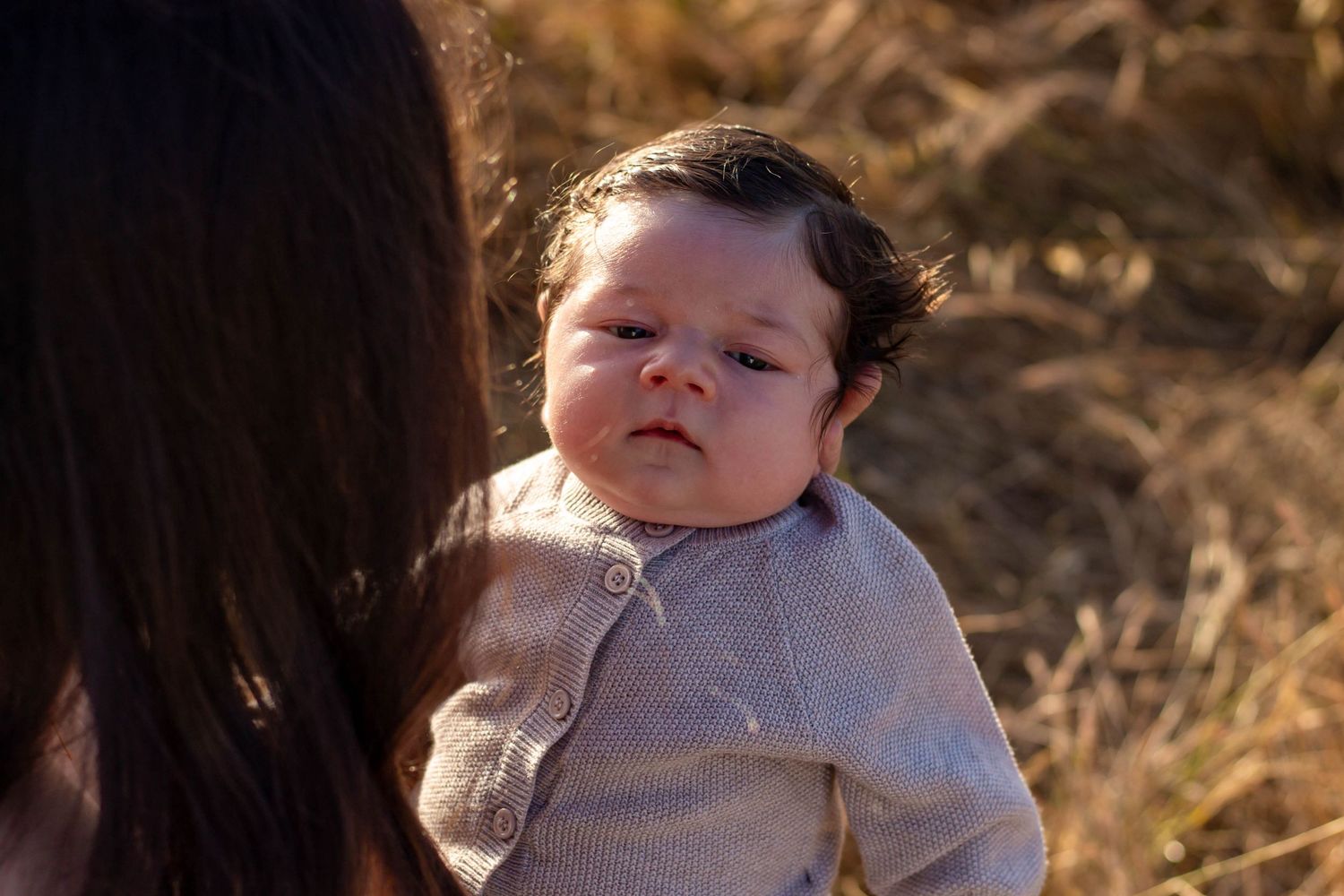 Close-up portrait of newborn baby with dark hair in gray cardigan held by mother outdoors.