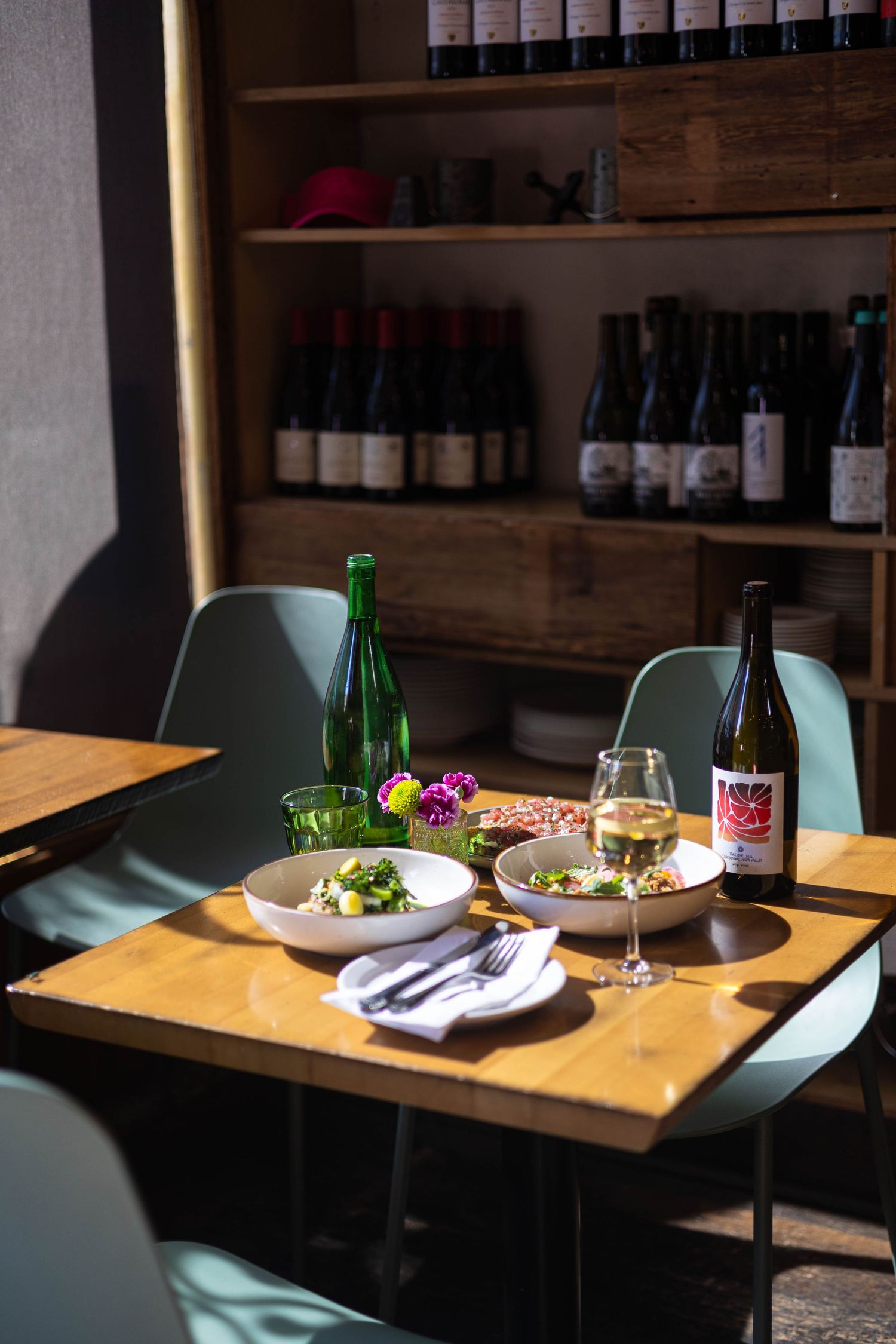 Sunlit restaurant table with two salad dishes, white wine glass, green bottle, and wine rack with bottles in background.