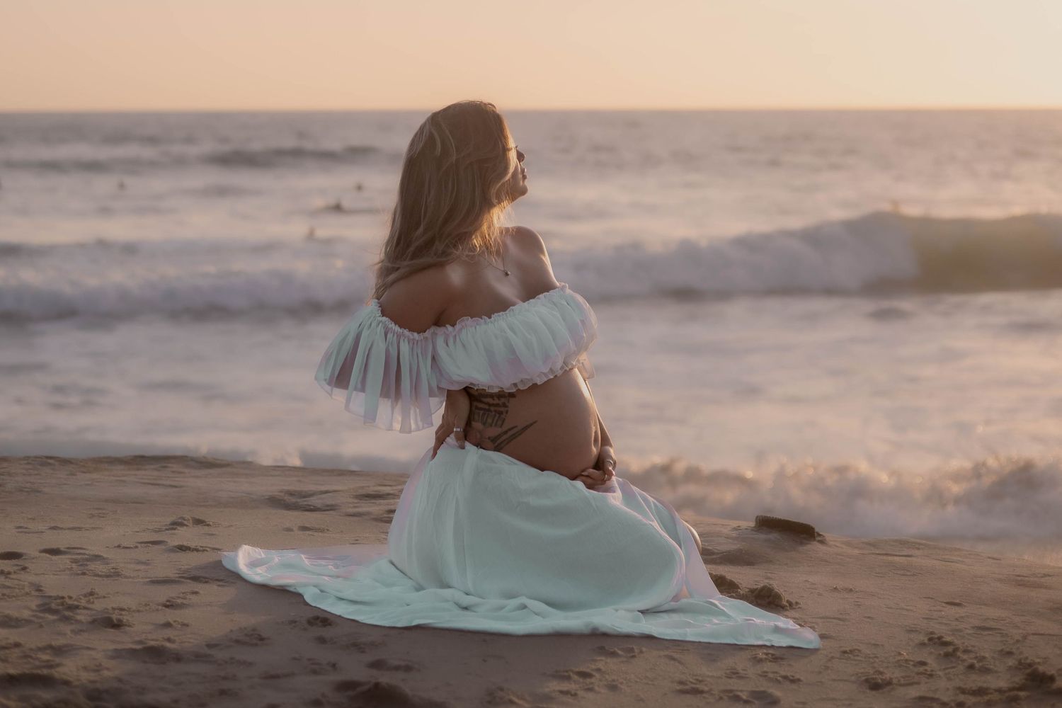 Pregnant woman in white off-shoulder dress kneeling on beach cradling bump at golden sunset.