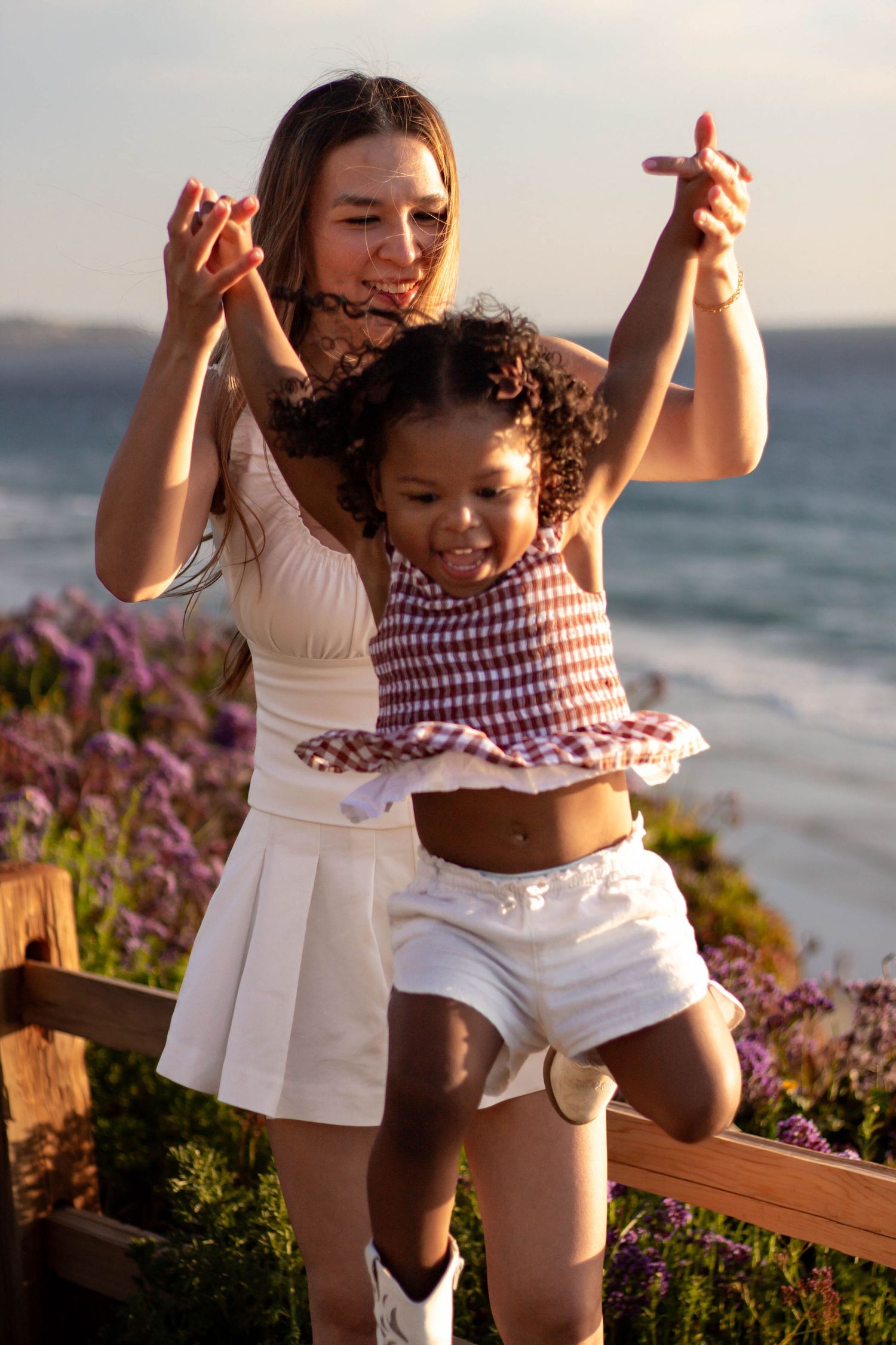 A smiling mother lifts her young daughter by the hands near a coastal garden at golden hour.