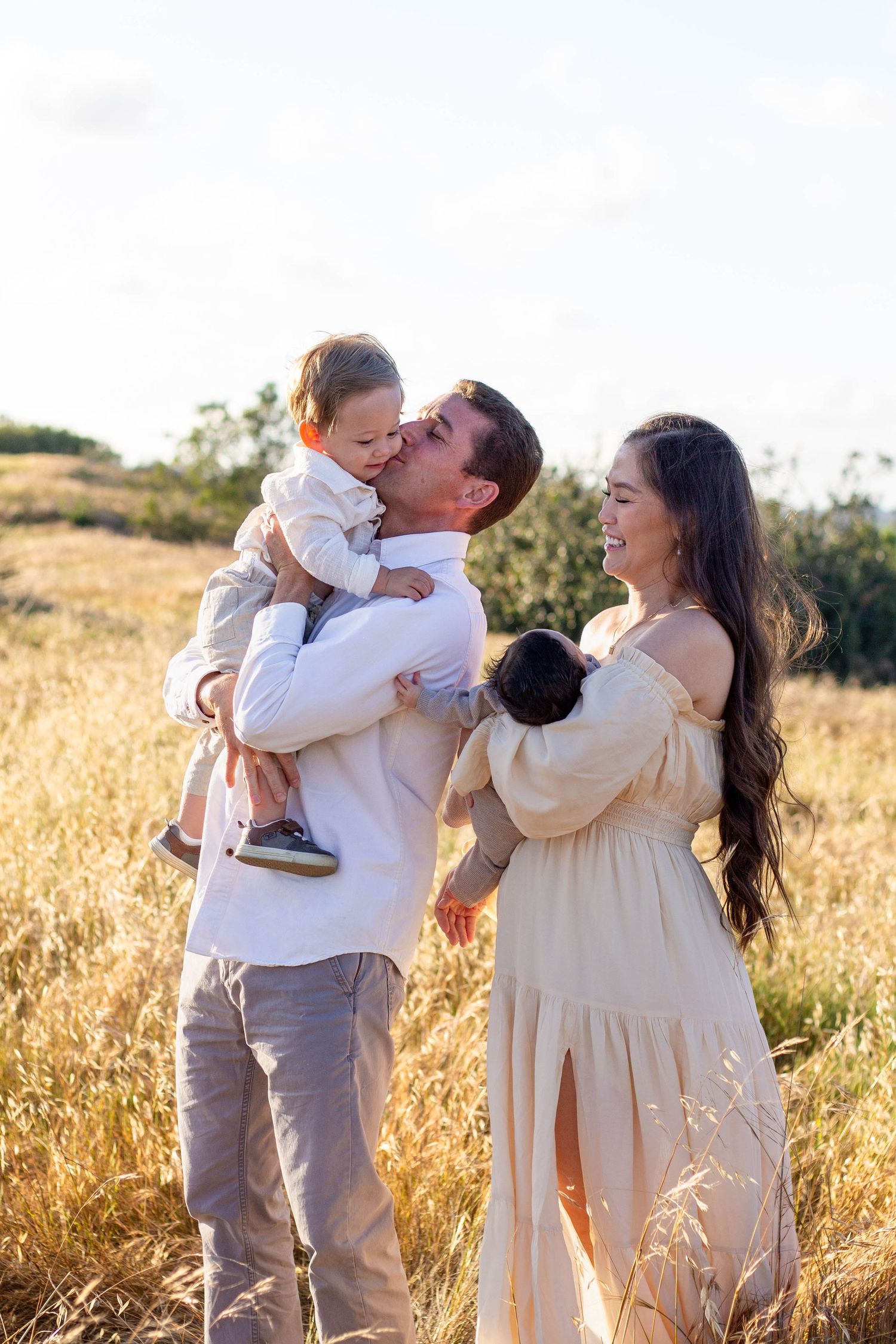 Happy family of four in golden field, father kissing toddler while mother holds newborn.