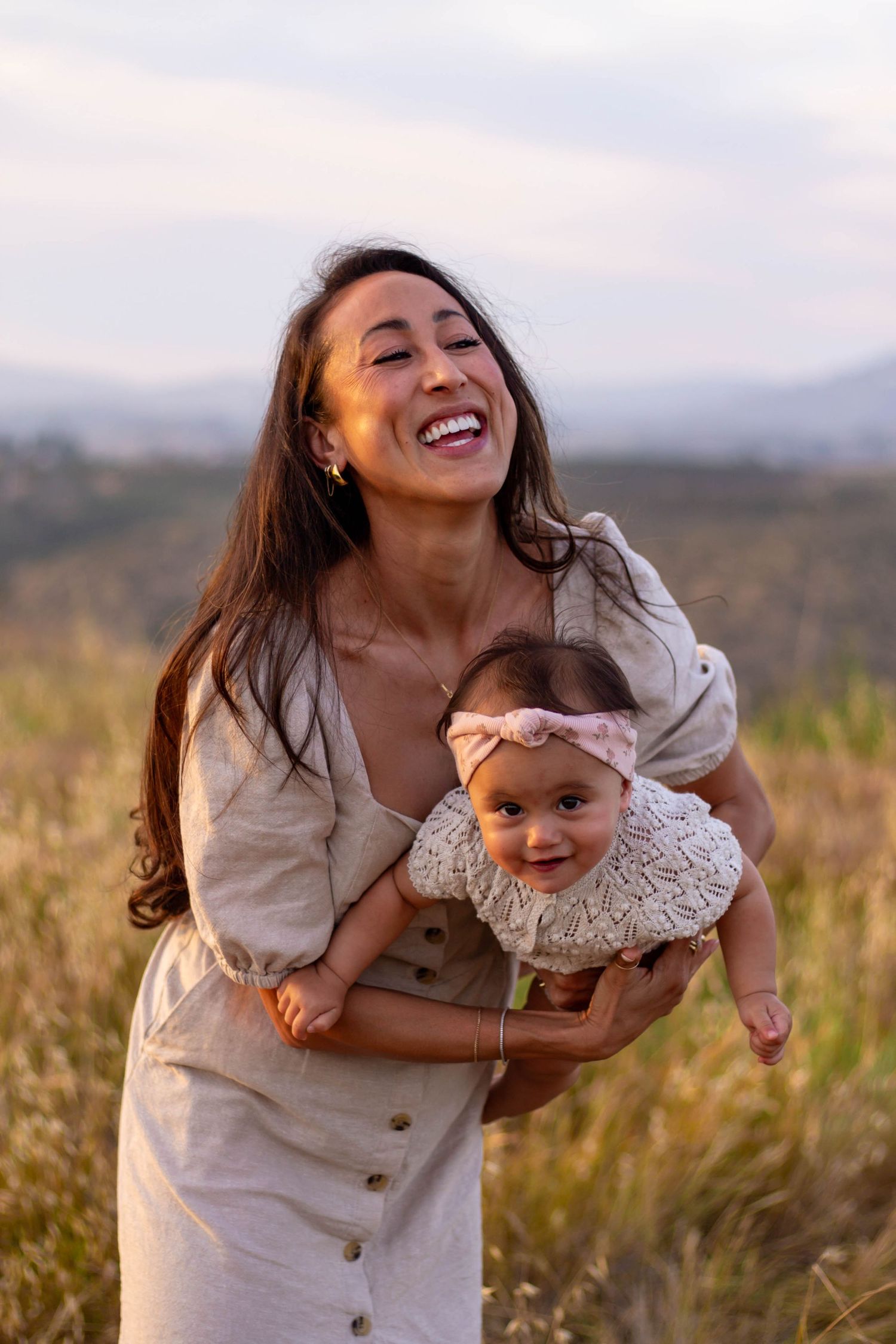 Joyful mother laughing while holding smiling baby girl in linen dress in golden field.