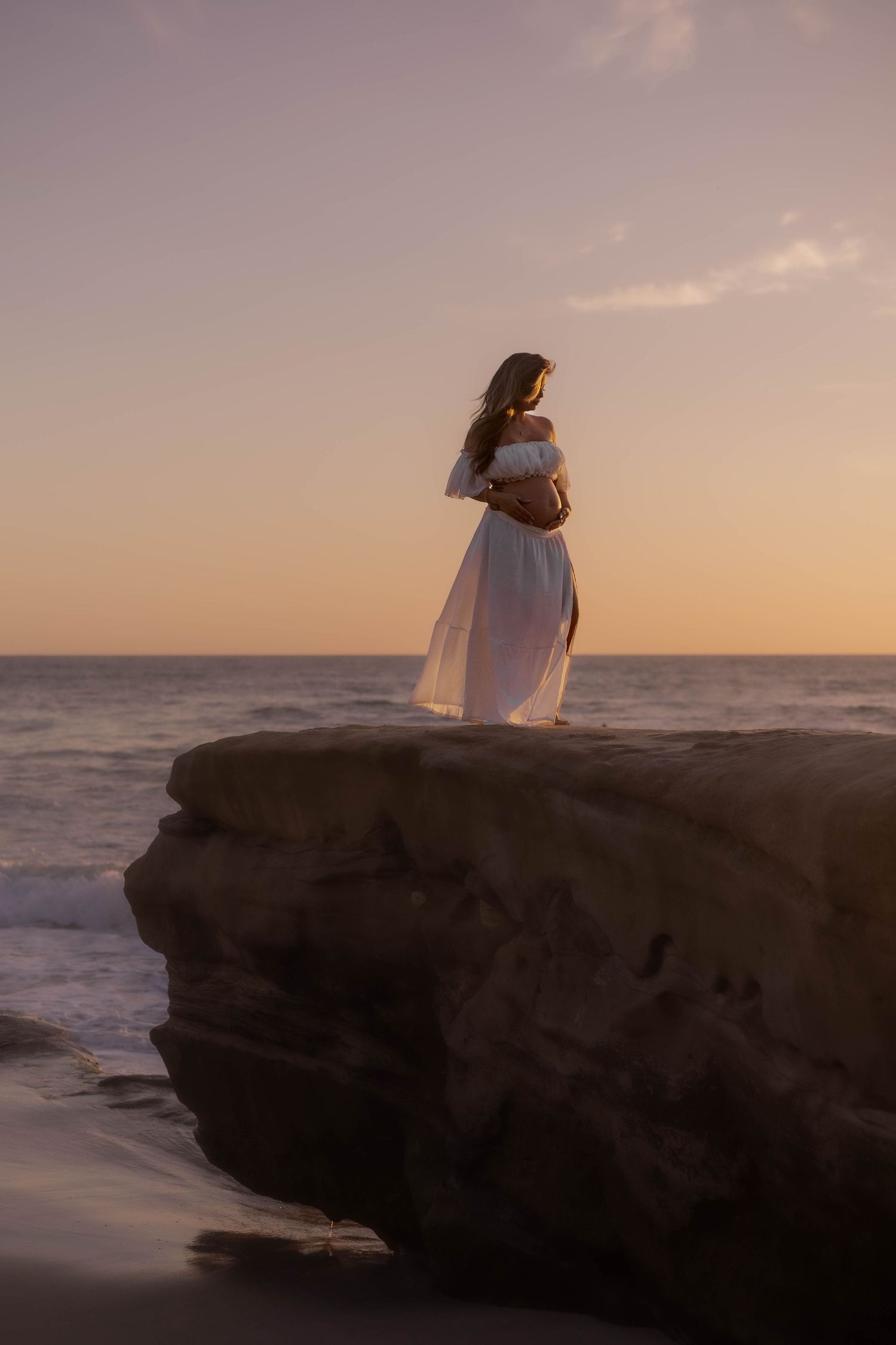 Pregnant woman in flowing white dress standing on coastal rocks at sunset.