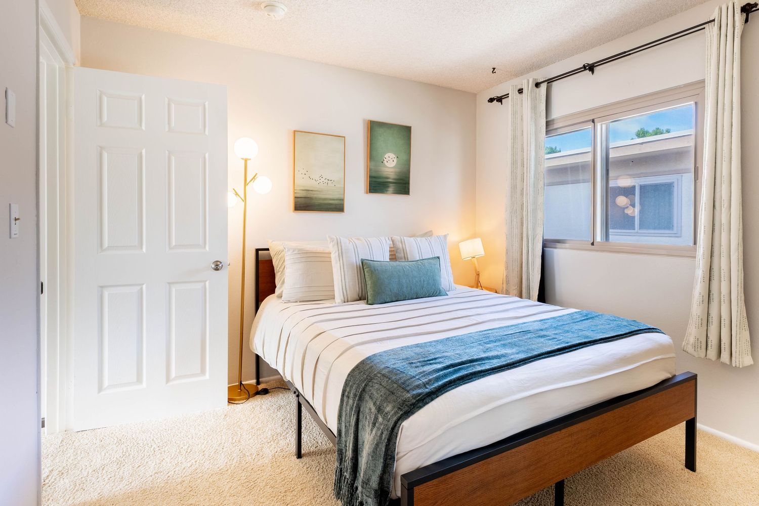 Cozy bedroom with a queen bed, blue accents, carpet flooring, white closet door, and a bright window with curtains.