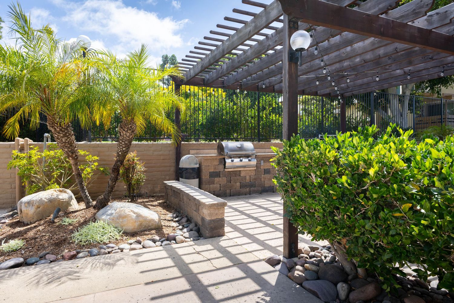 Outdoor patio with pergola, BBQ grills, lush tropical plants, and decorative rocks in a sunny landscaped setting.