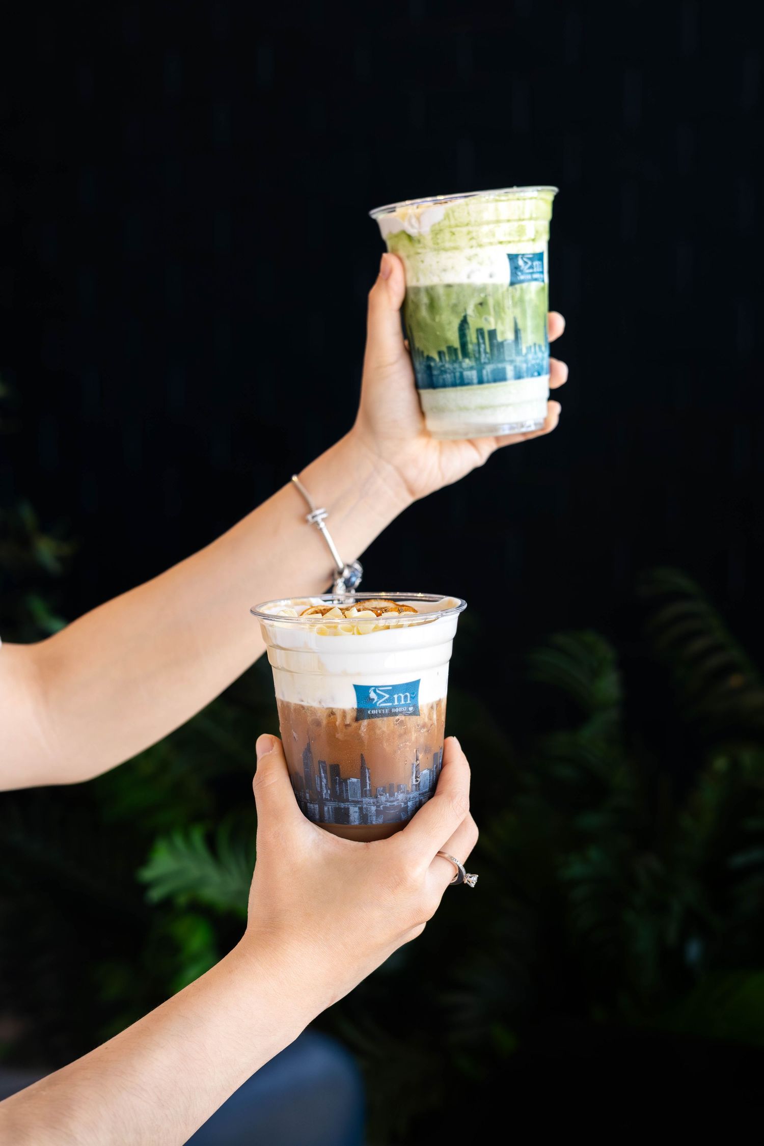 Two hands holding layered iced drinks in branded cups with matcha and coffee flavors against a dark background.