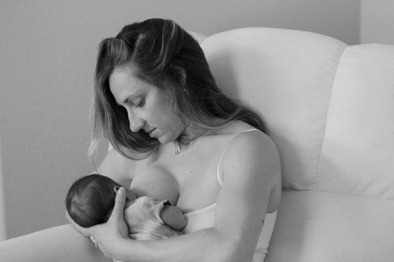 Black and white photo of a mother tenderly cradling and breastfeeding her newborn baby while seated in a chair.