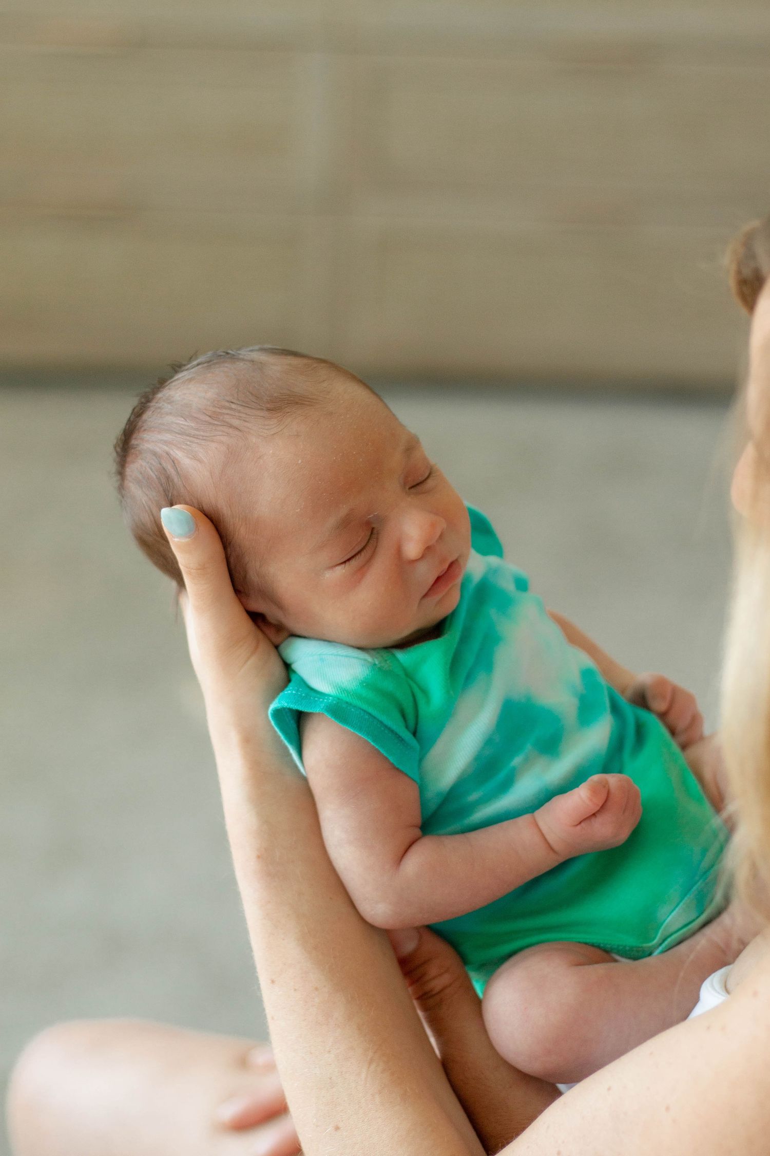 A newborn baby in a green onesie sleeping peacefully while being cradled in an adult's hands.