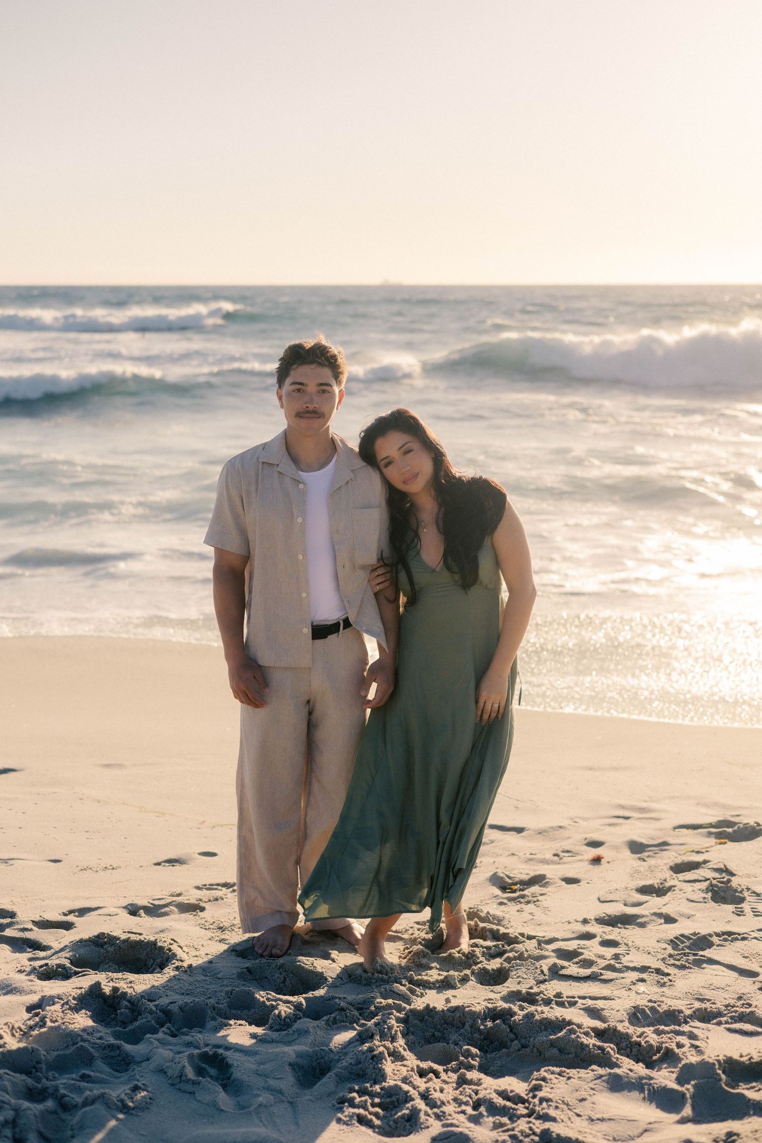 Young couple standing together on beach at sunset, woman in green dress leaning on mans shoulder.