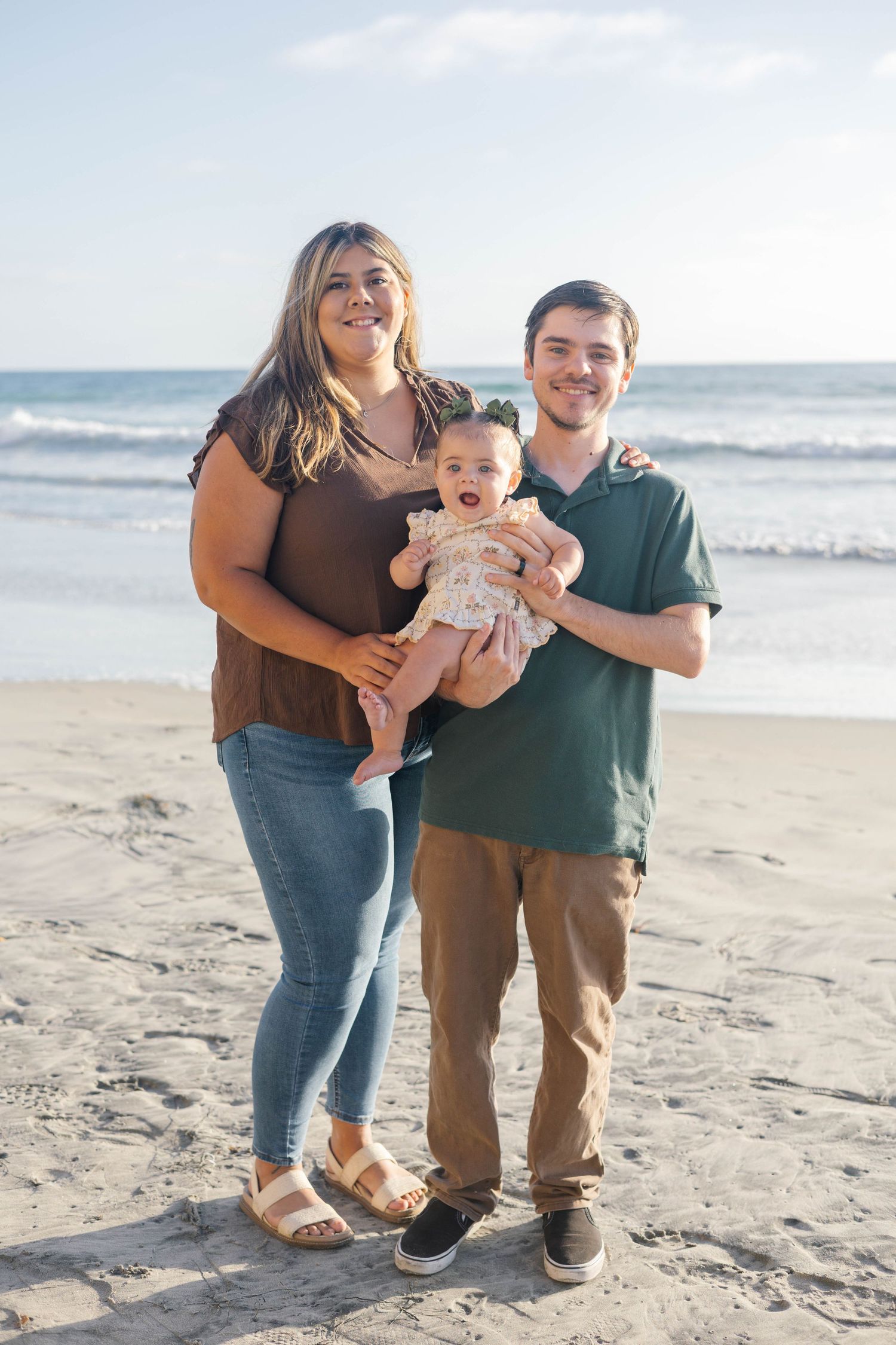 A young family of three poses on a sandy beach, with a woman and man smiling while holding their happy baby girl.