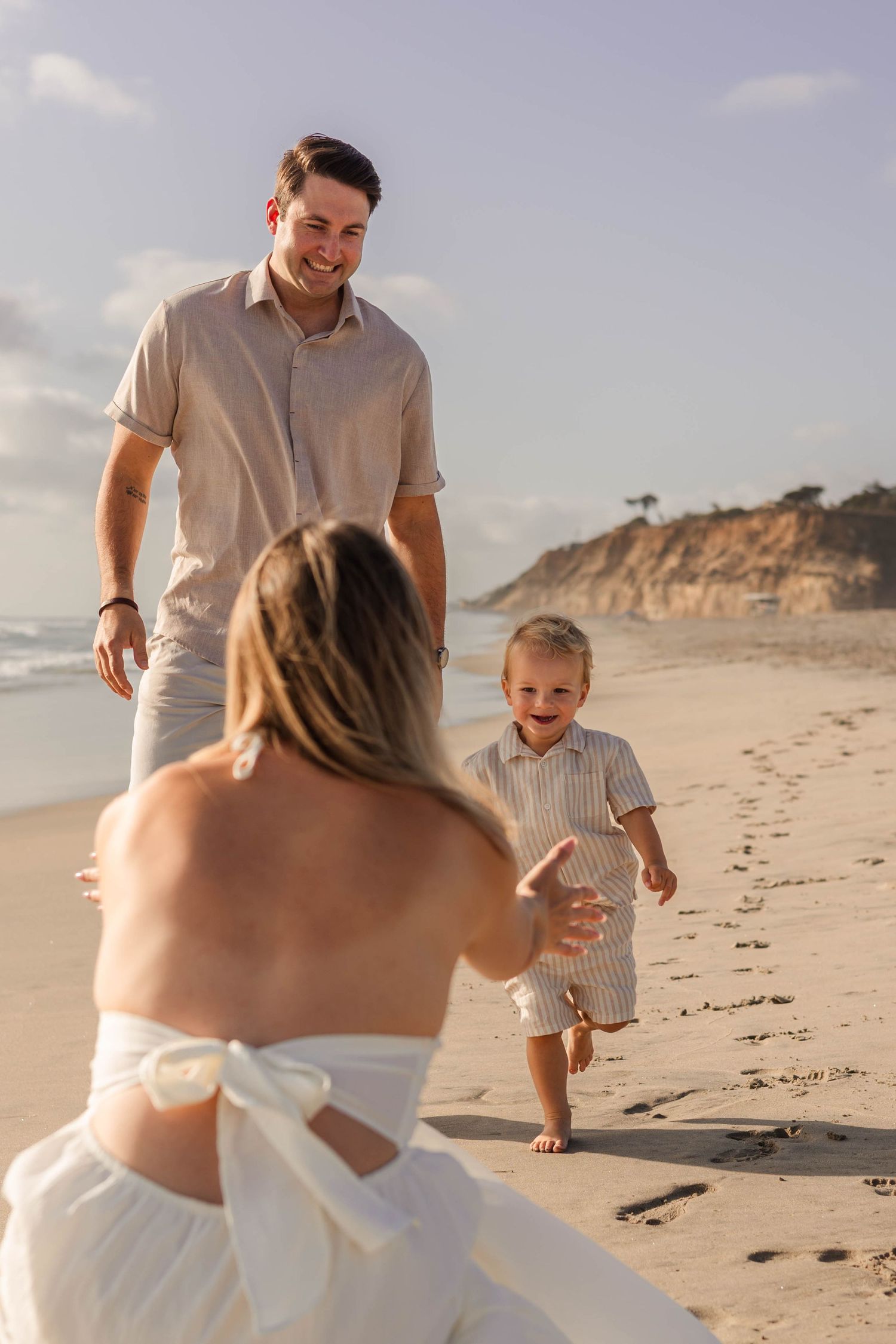A joyful toddler runs toward his mother on a sunny beach while his smiling father watches nearby.