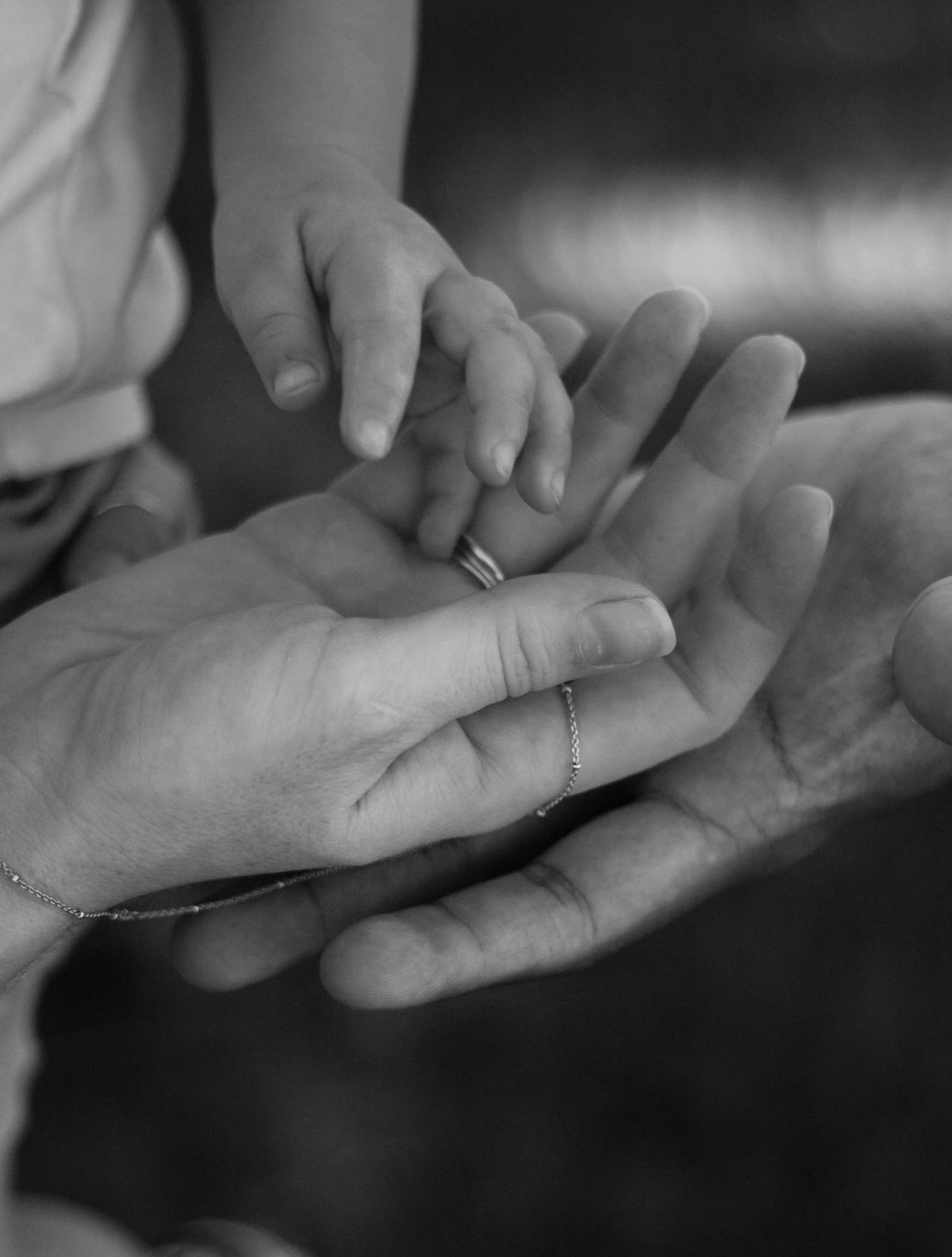 A black and white close-up of a baby's tiny hand resting on two adult hands with rings.