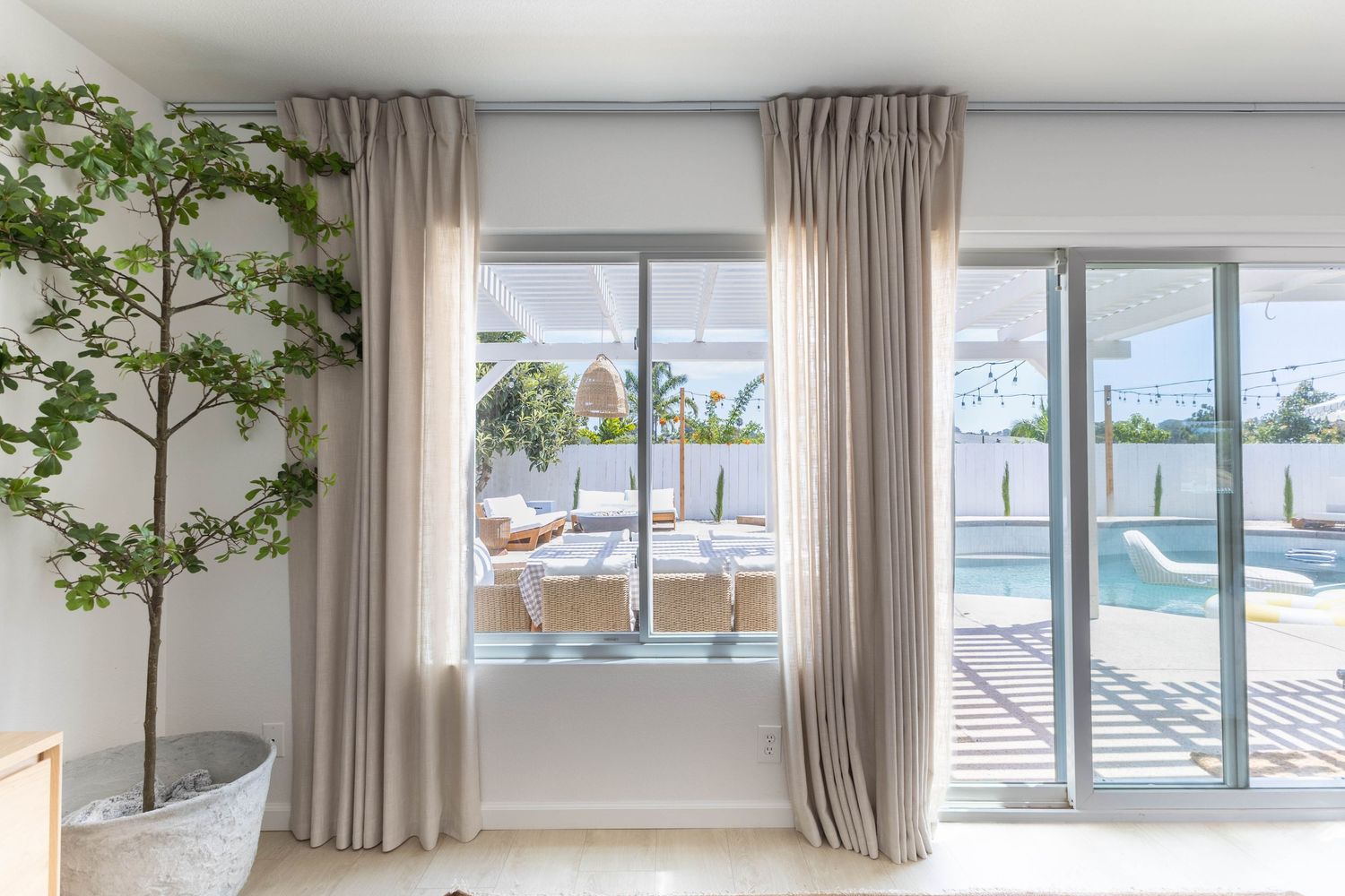 Elegant living room with linen curtains framing sliding glass doors overlooking a sunny outdoor pool patio area.