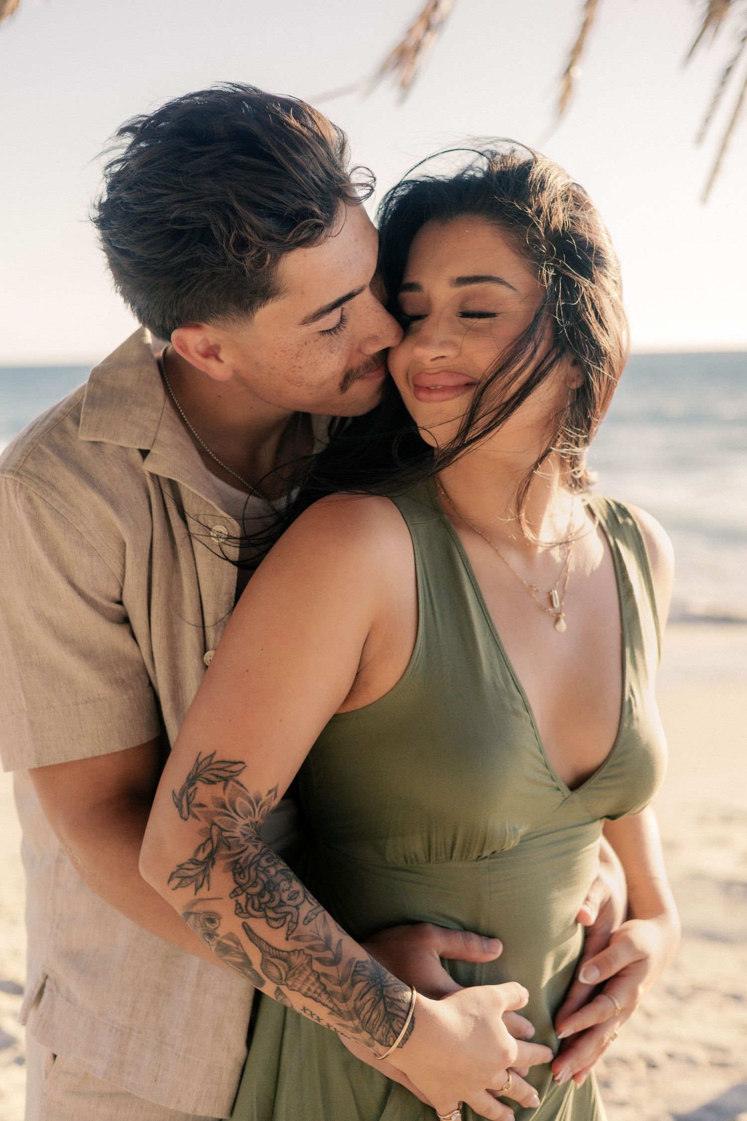 Man kissing womans cheek from behind while embracing her on beach at golden sunset hour.