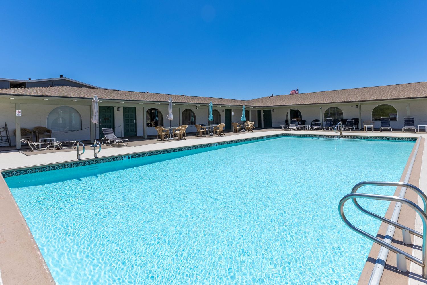Large outdoor swimming pool with clear blue water surrounded by lounge chairs and a motel building with green doors.