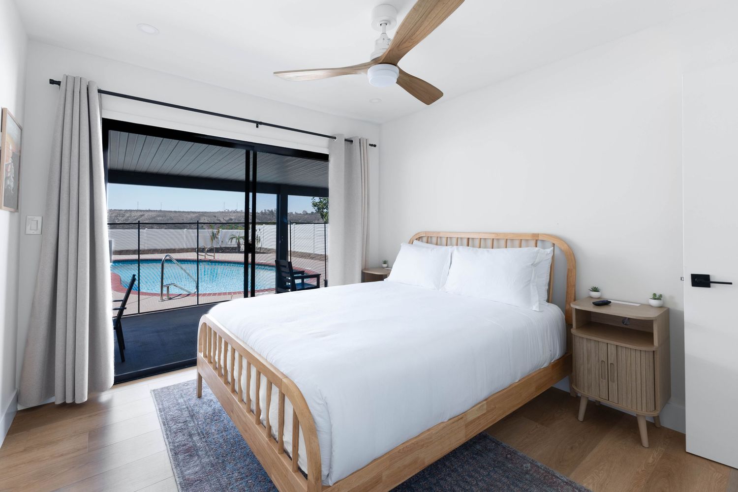 Bright bedroom with rattan bed, white bedding, hardwood floors, ceiling fan, and sliding doors opening to a pool view.